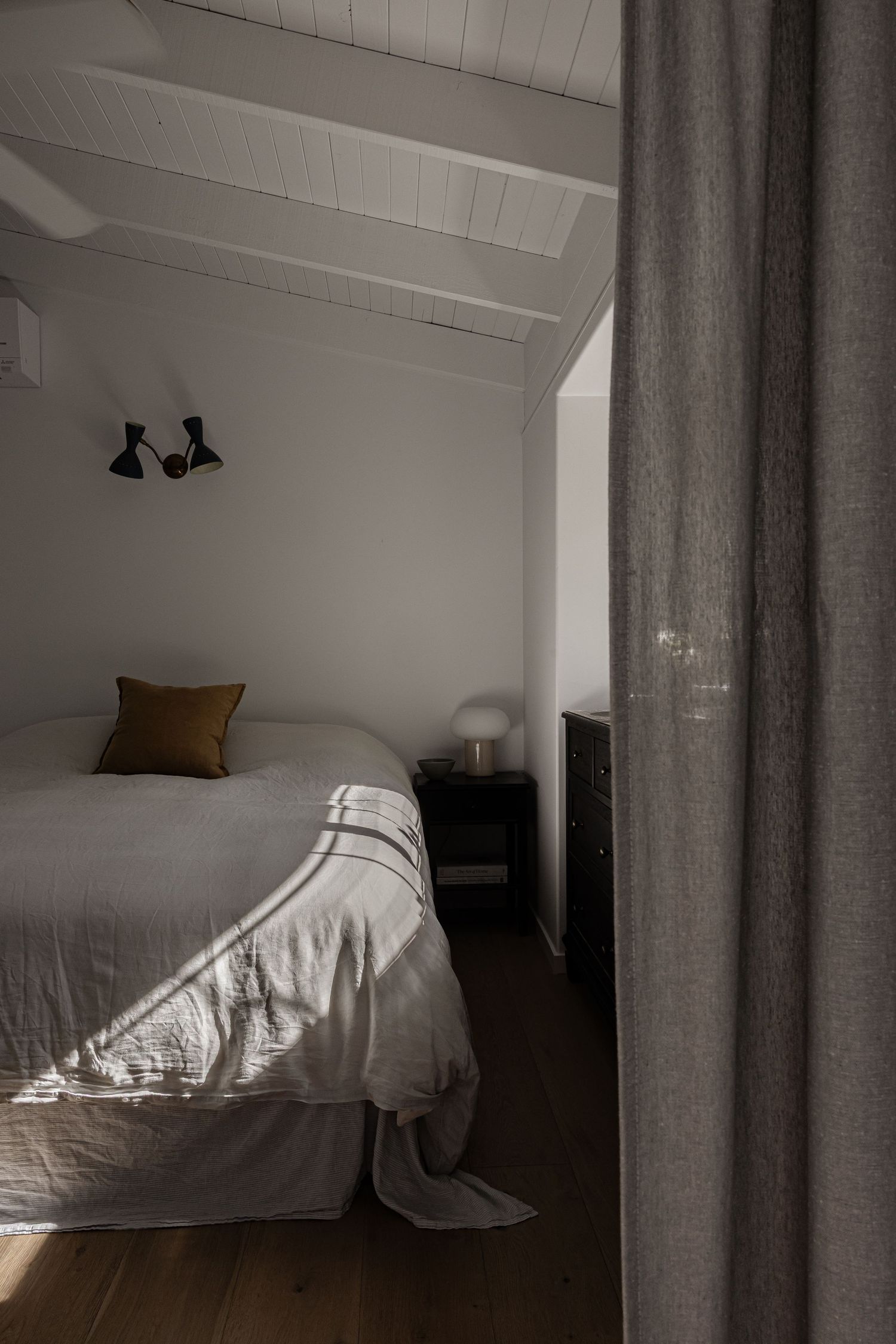A minimalist bedroom interior with white bedding, brown hardwood floors, and black wall sconces in natural daylight.