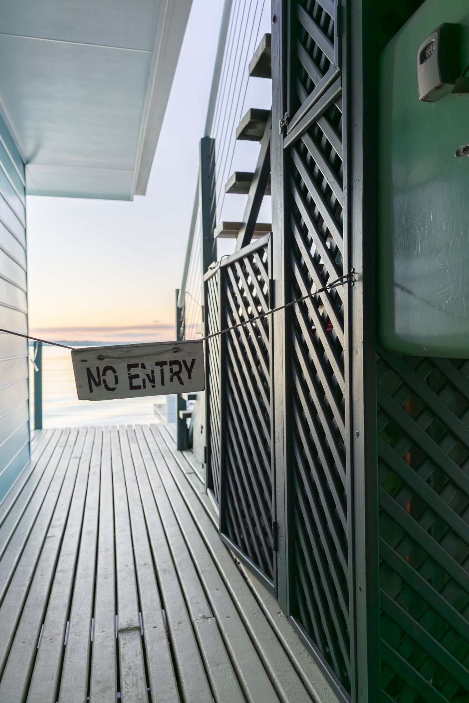 Wooden boardwalk with 'No Entry' sign and lattice railings overlooking ocean at sunset.