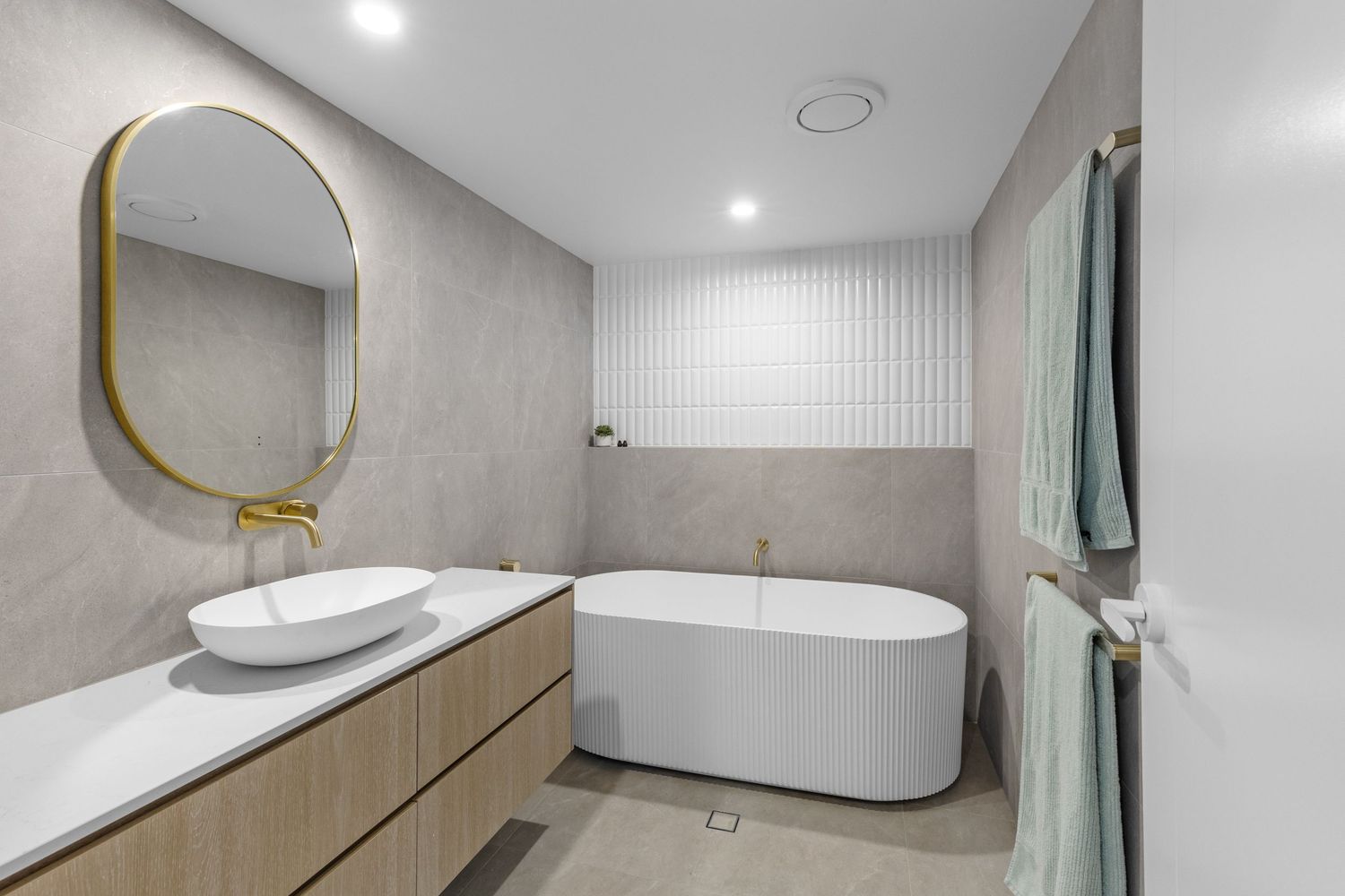 Modern bathroom interior with freestanding oval tub, wooden vanity, and gold-framed round mirror against gray walls.