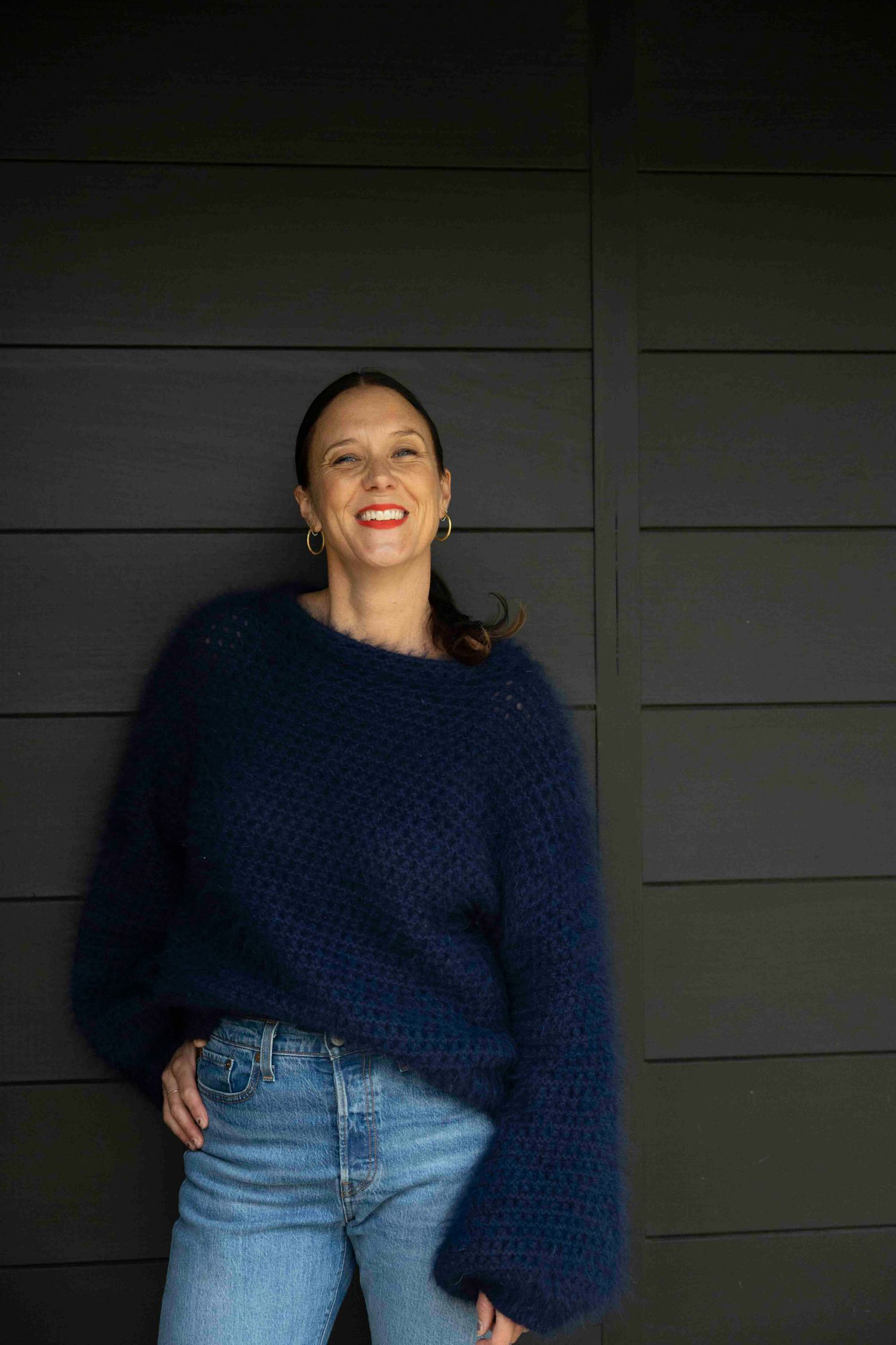 Person wearing navy knit sweater and jeans stands against dark wall background.