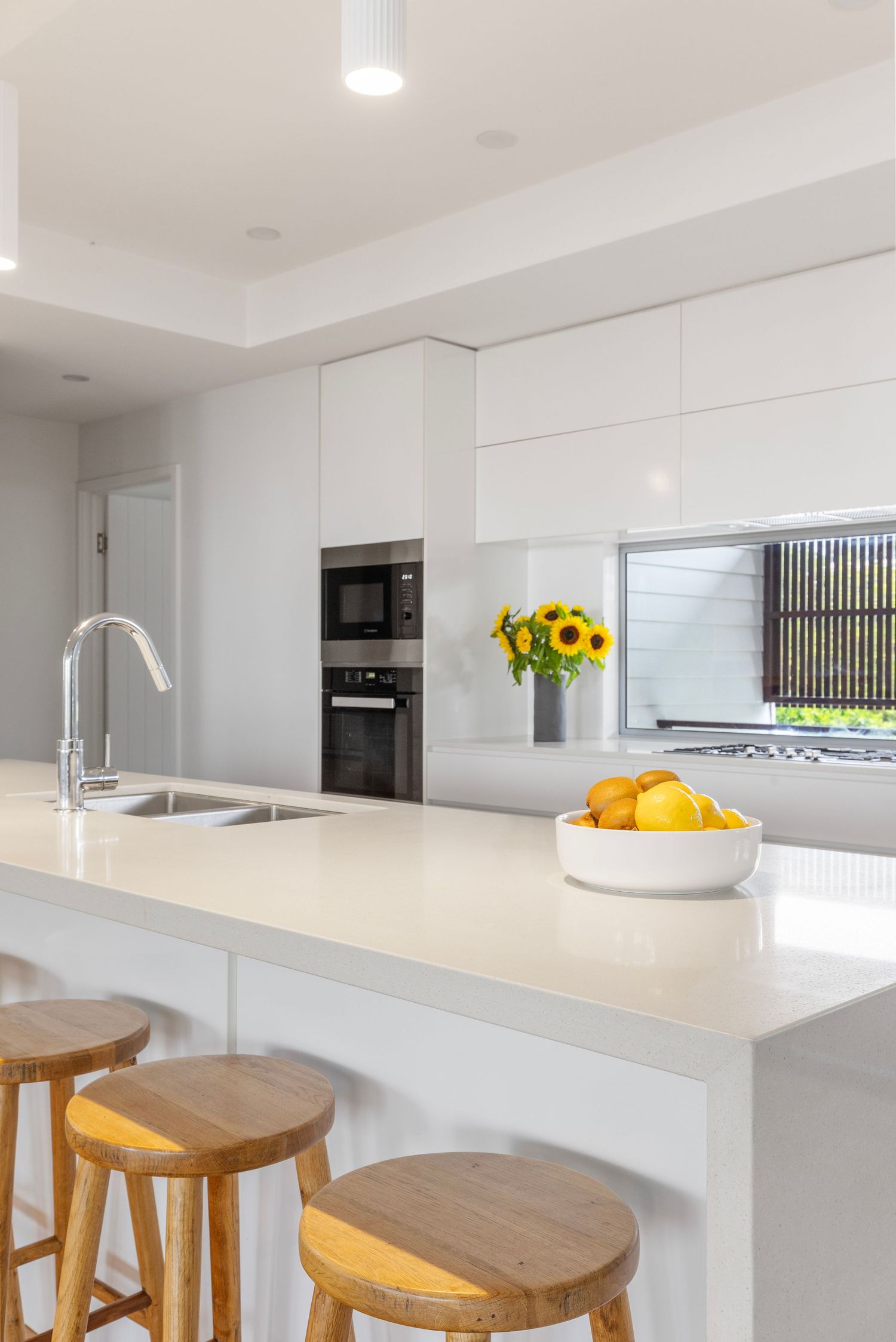 Modern white kitchen with sleek countertops, wooden bar stools, and bright yellow sunflowers in a vase by the window.