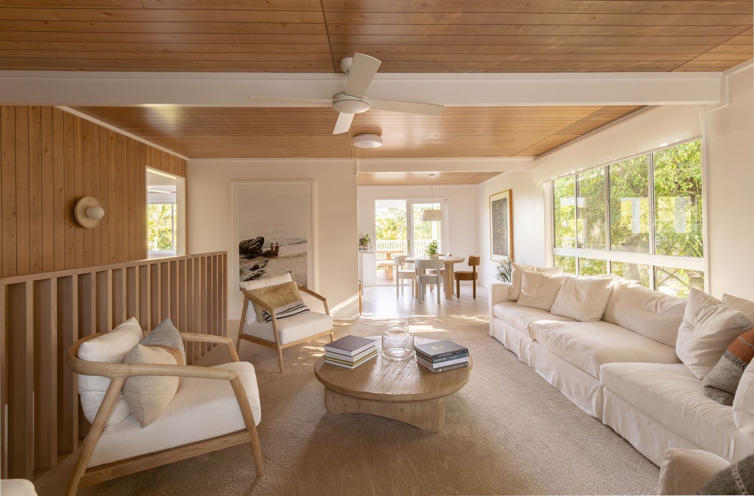 Bright and airy coastal living room with white furniture, wood paneling, and large windows overlooking tropical greenery.