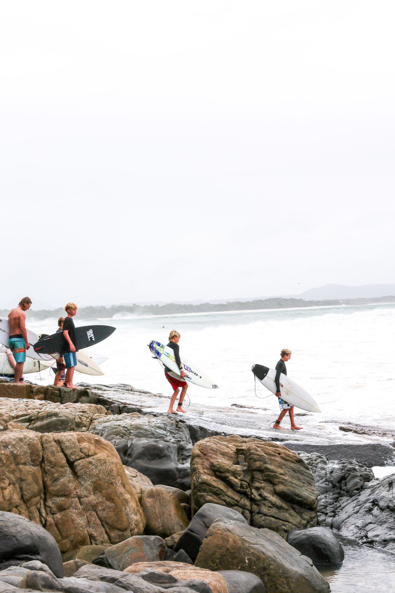 Surfers carrying boards traverse rocky coastline during overcast day.