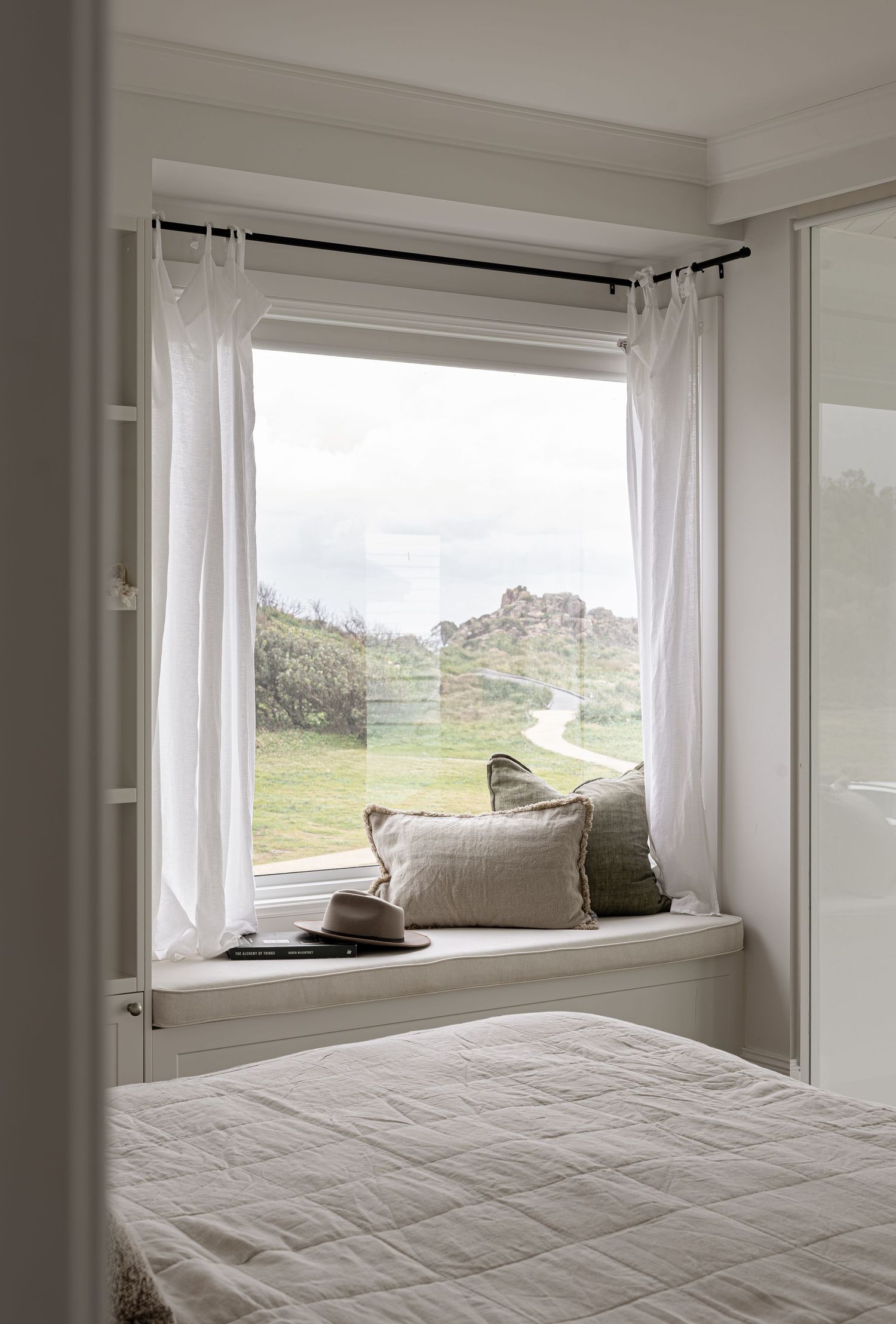 A cozy window seat with white curtains and cushions overlooks a green hillside landscape, creating a serene bedroom nook.