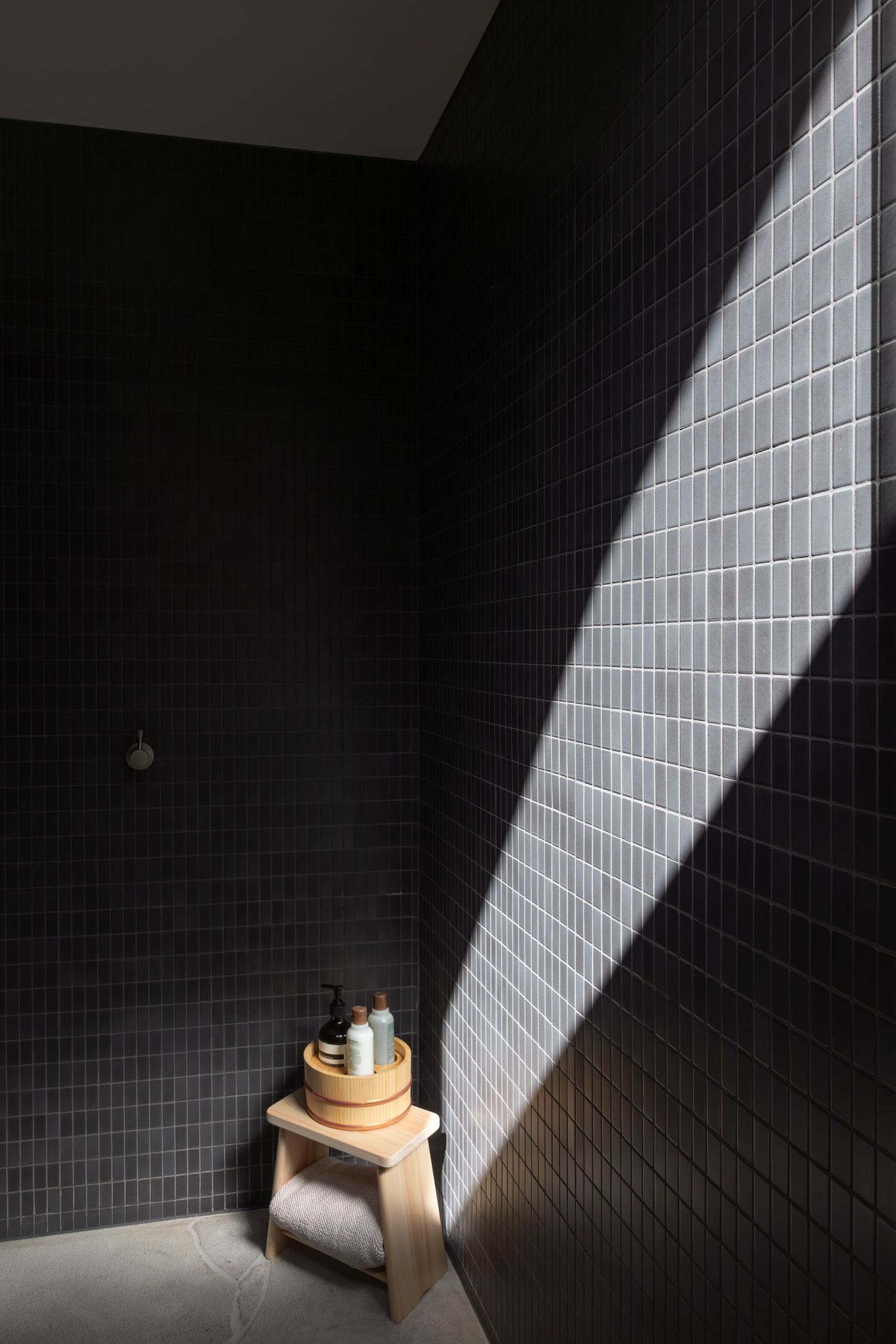 A dramatic beam of natural light illuminates a wooden stool against dark tiled walls in a minimalist bathroom setting.