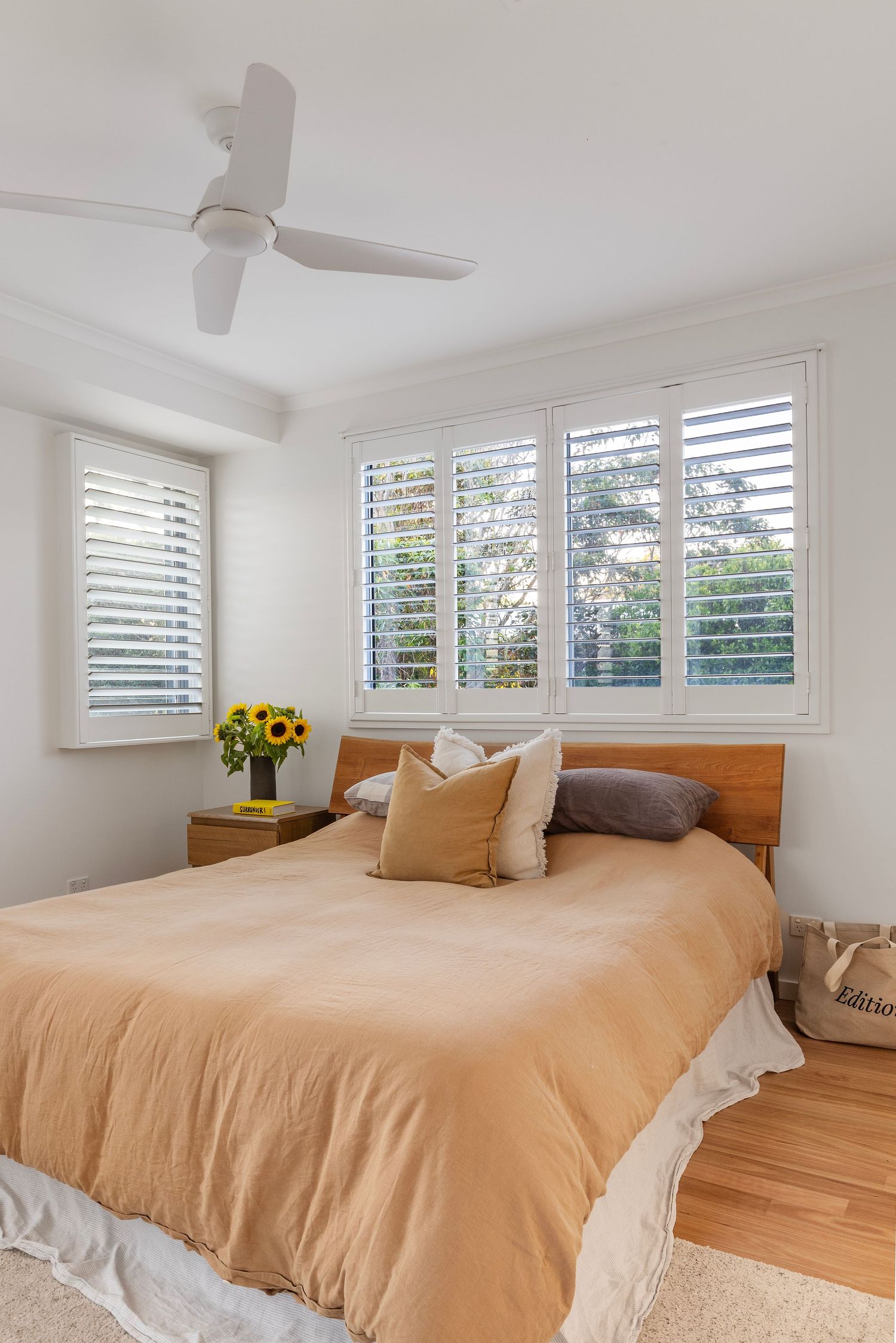 Modern white bedroom with plantation shutters and peach-colored bedding features a row of windows letting in natural light.