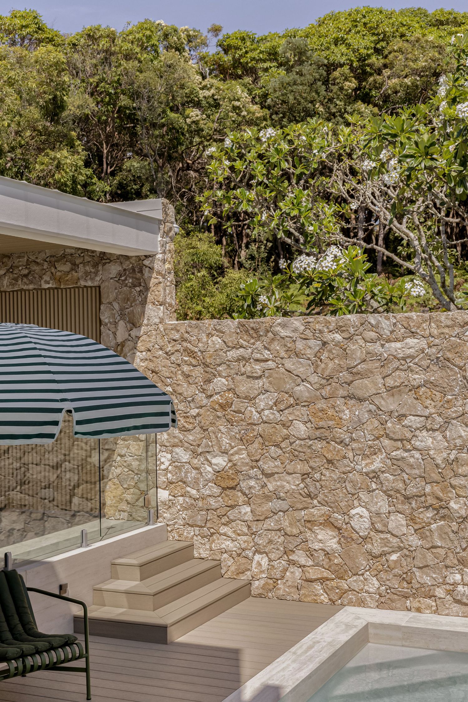 Stone-walled outdoor pool area with timber decking, glass balustrade, striped umbrella, and lush green trees backdrop.