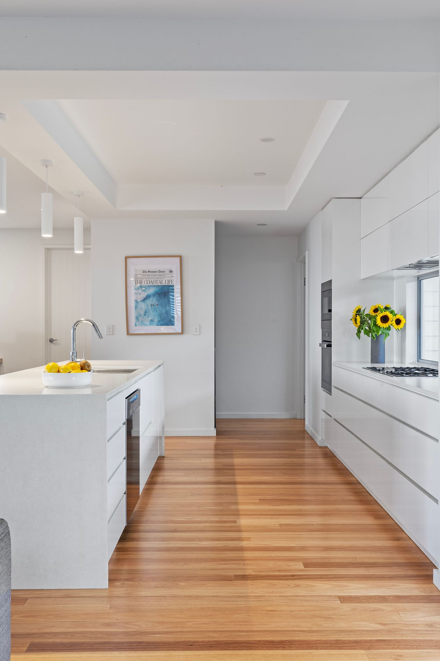 Modern white kitchen with long galley layout features sleek cabinets and warm hardwood floors extending down hallway.