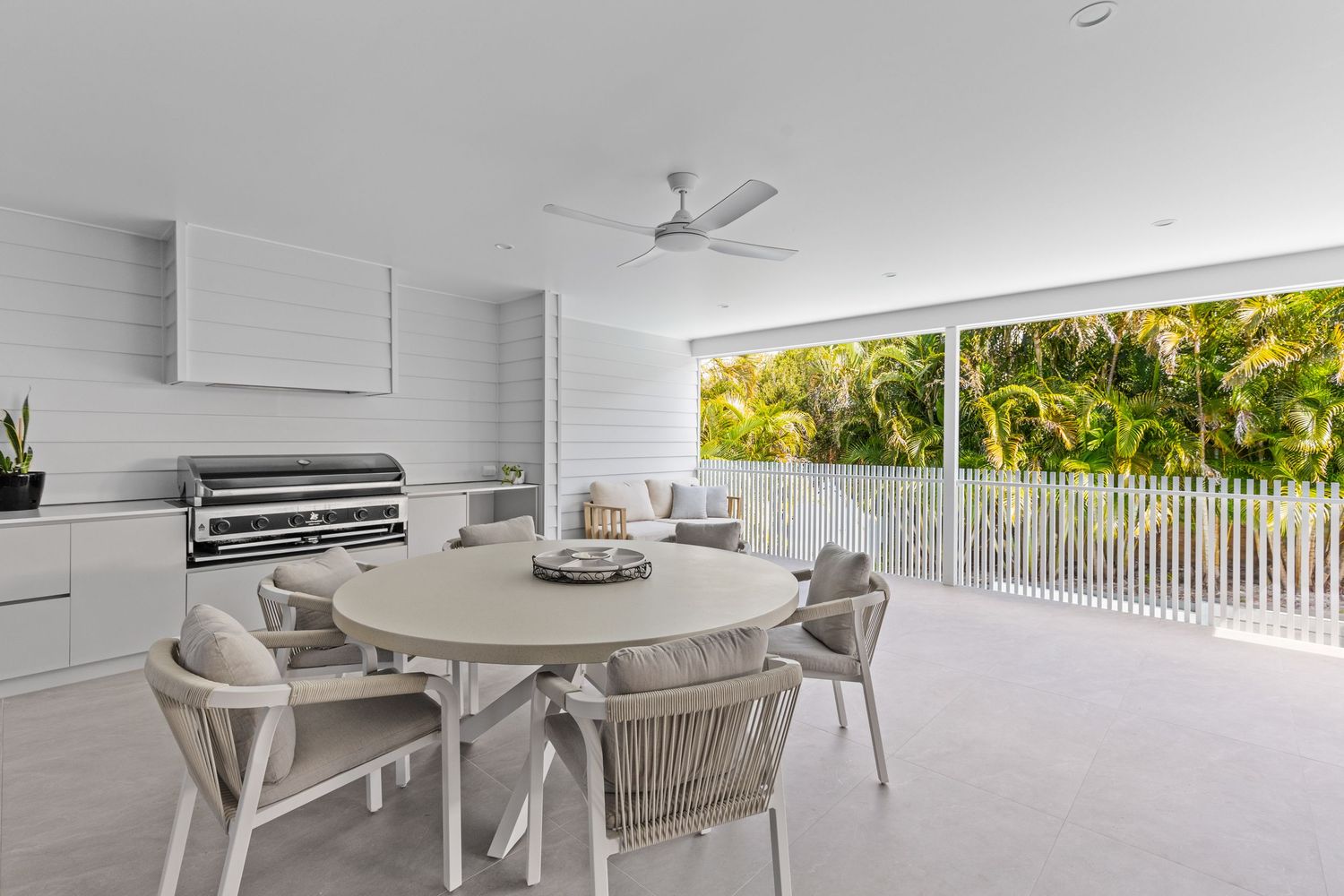 Modern covered outdoor patio with round dining table, gray chairs and tropical garden views through white railings.