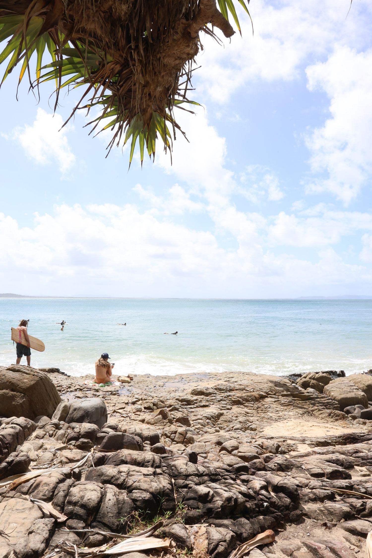 Beachgoers enjoy sunny day among coastal rocks with palm fronds overhead.
