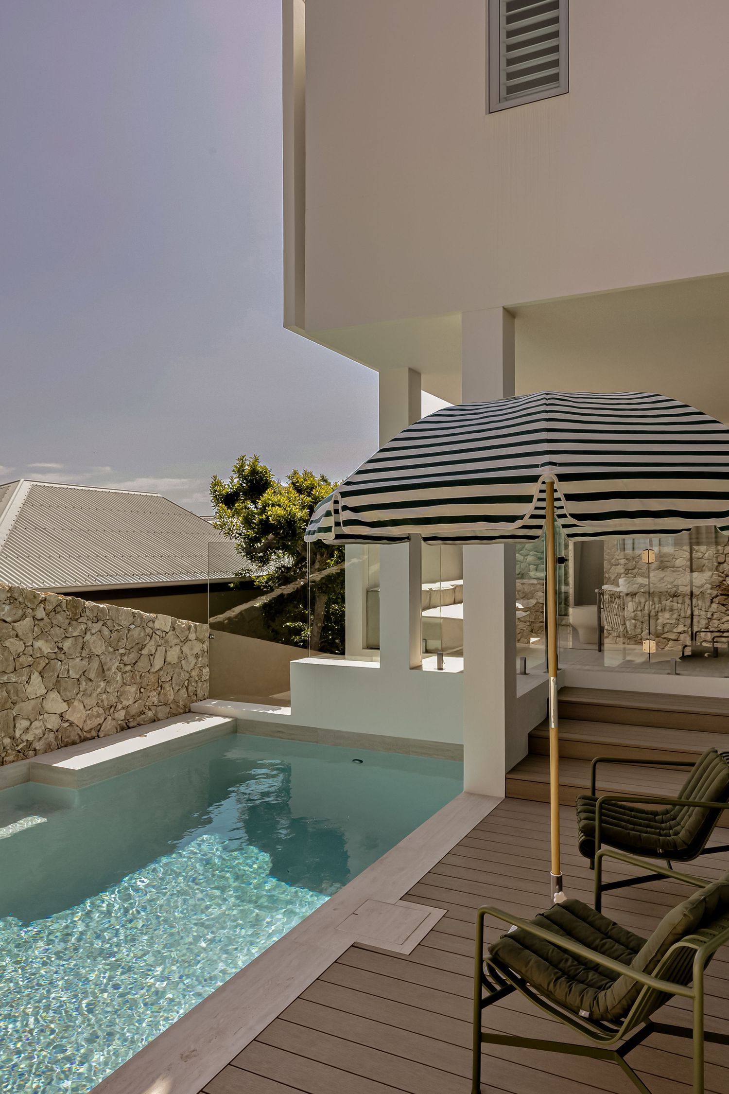 Modern white villa with a turquoise plunge pool, striped umbrella, and green lounge chairs on a wooden deck.