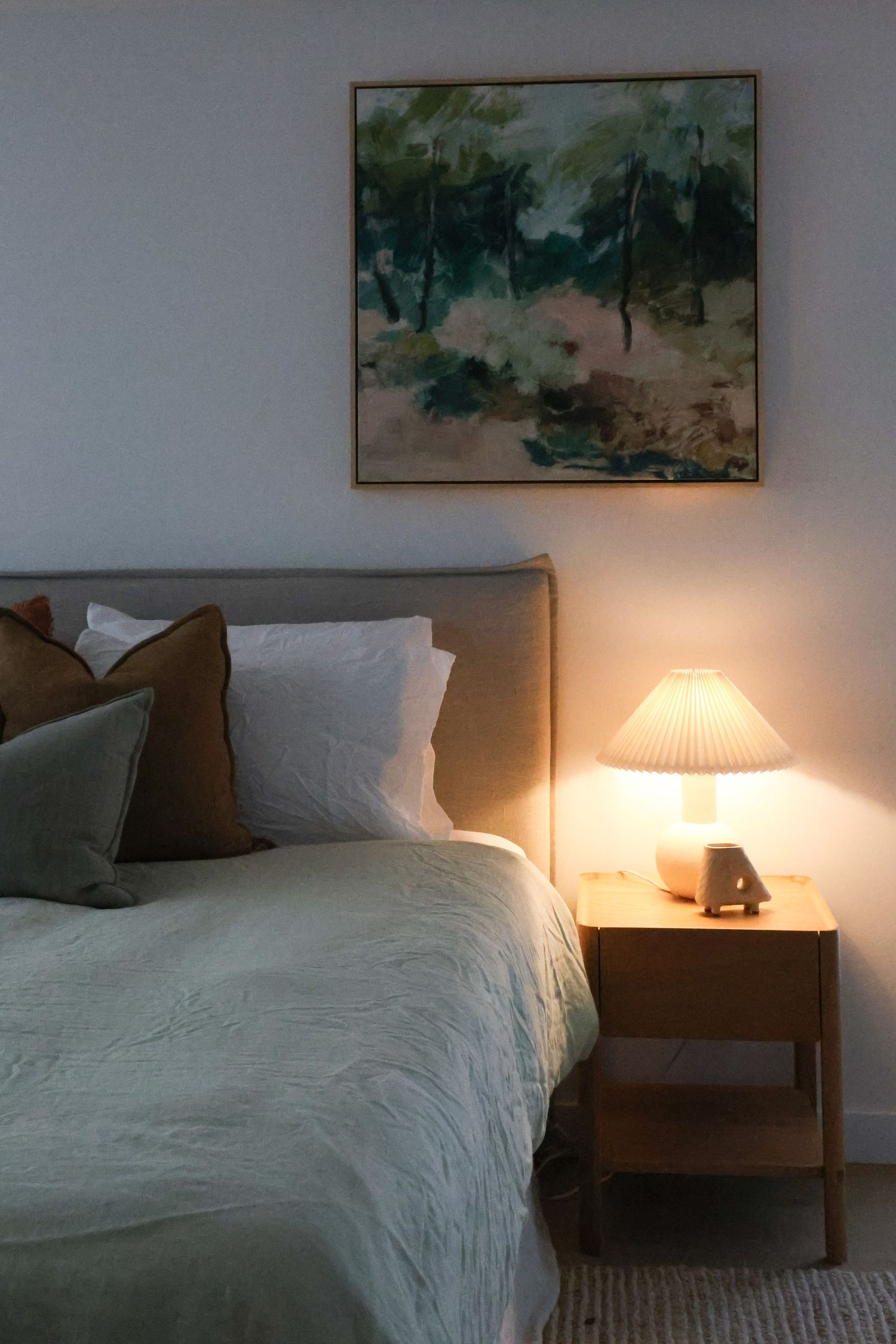 A bed with white linens and bedside lamp creating warm lighting in a cozy bedroom interior with artwork on the wall.
