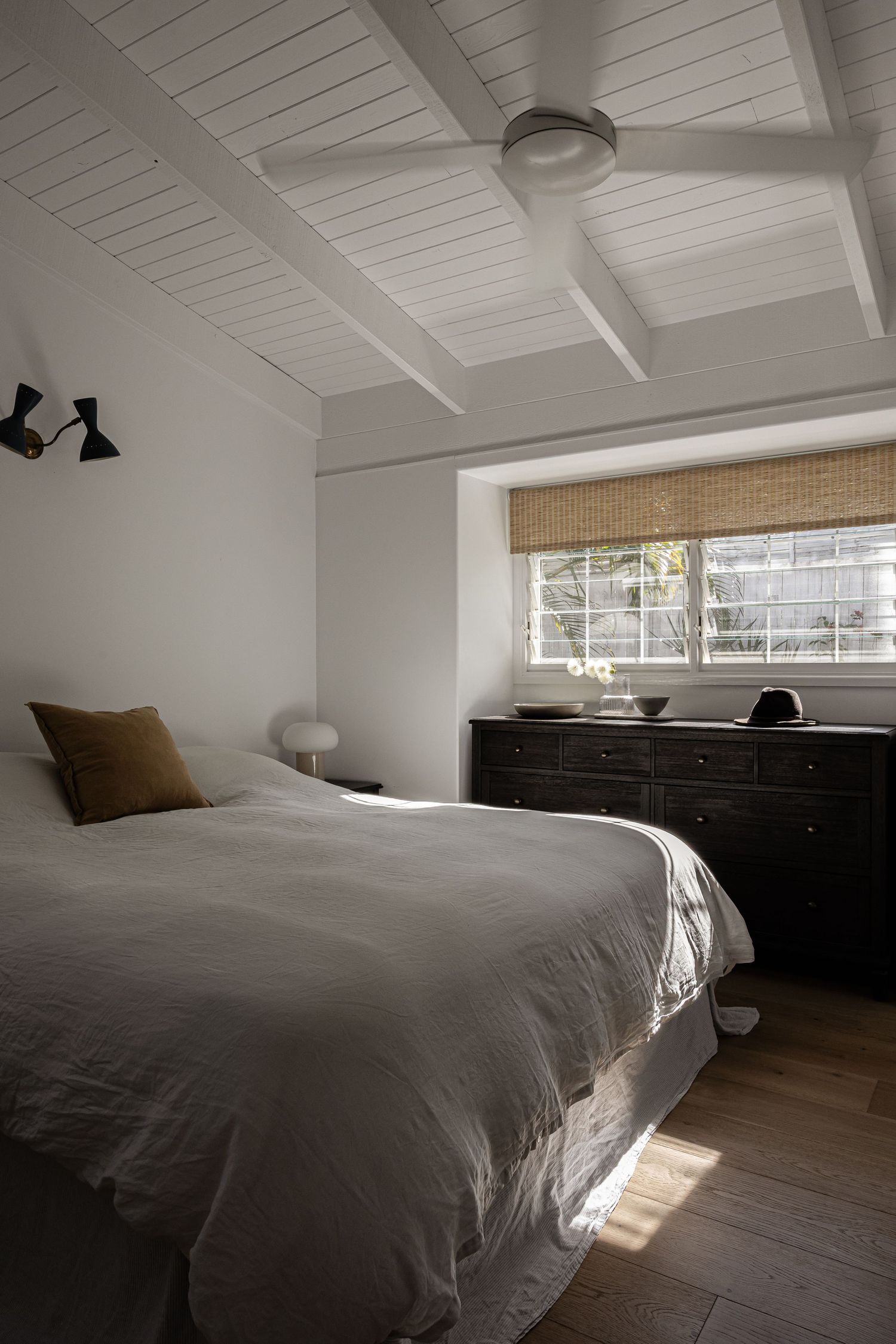 Modern minimalist bedroom with white ceiling, ceiling fan, bamboo shade, dark dresser and gray bedding on hardwood floors.