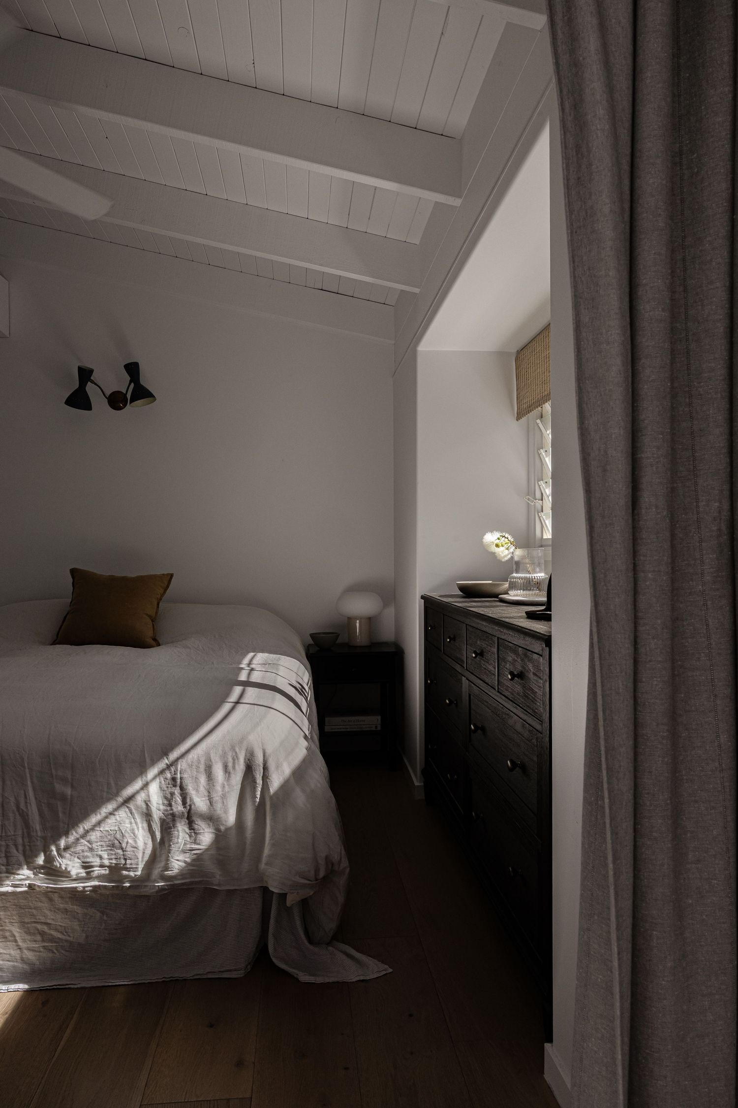 A modern minimalist bedroom at night features a white bed, gray curtains, and wooden flooring illuminated by soft lighting.