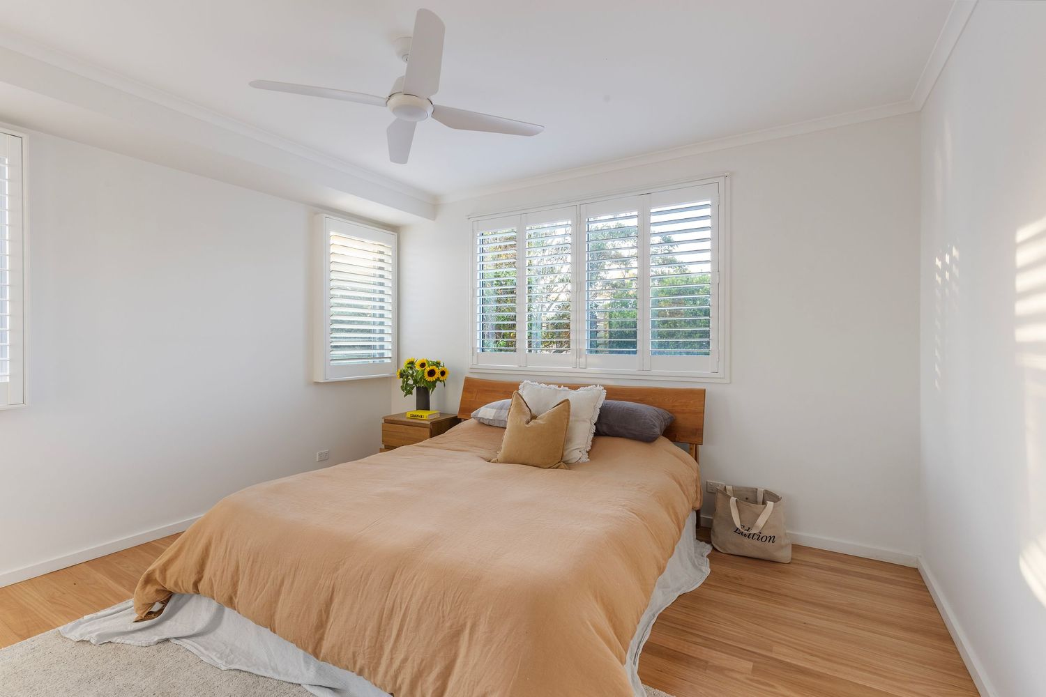 Light and airy bedroom interior with tan bedding, decorative pillows, hardwood floors and plantation shuttered windows.