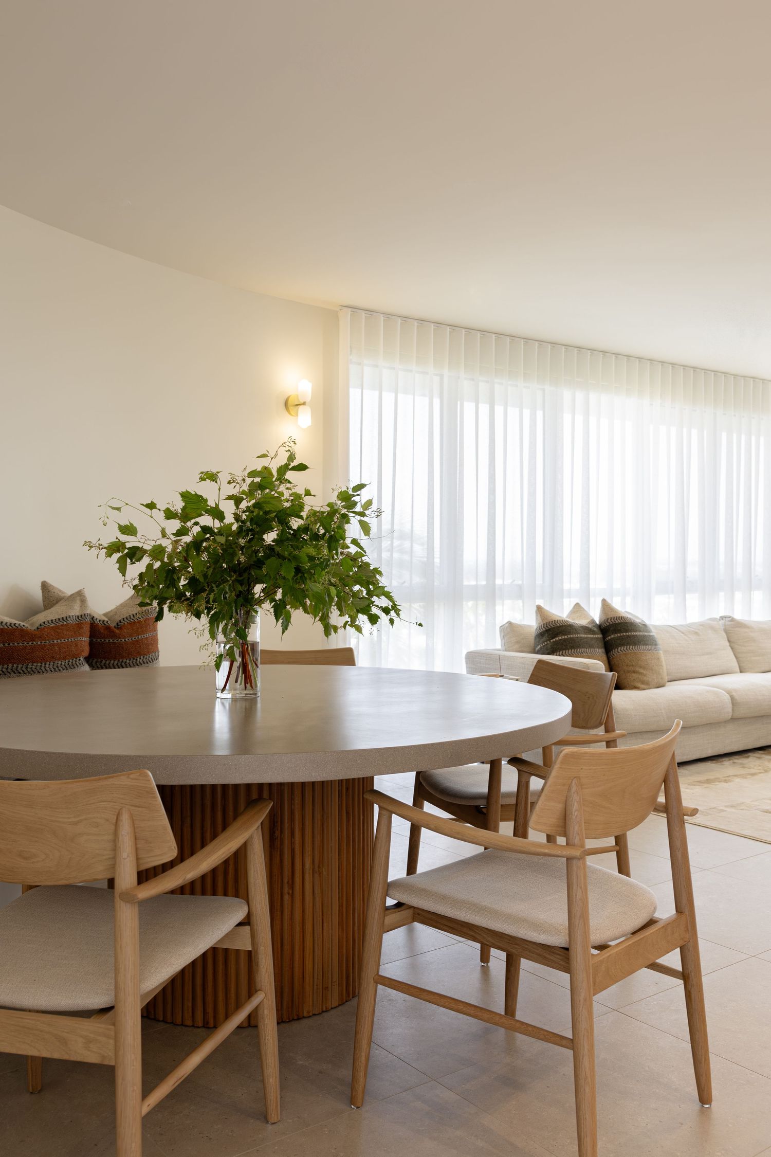 Minimalist dining area with round wooden table, light chairs, and potted plant against sheer white curtains in bright room.