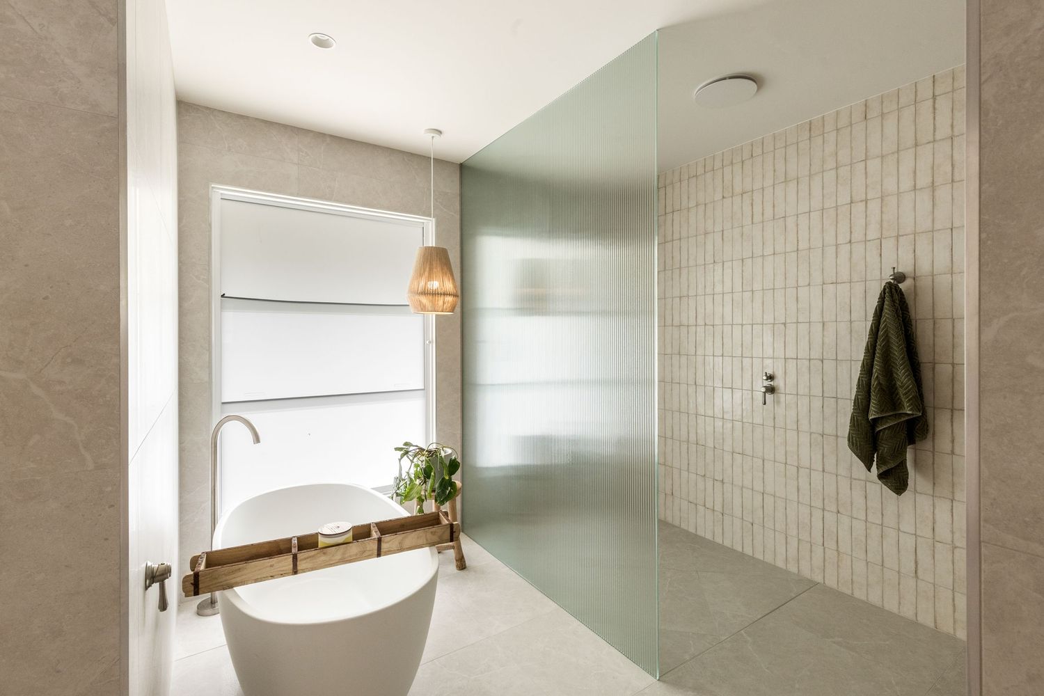 Modern bathroom interior featuring a freestanding white bathtub, beige tile walls, and natural light from a window.