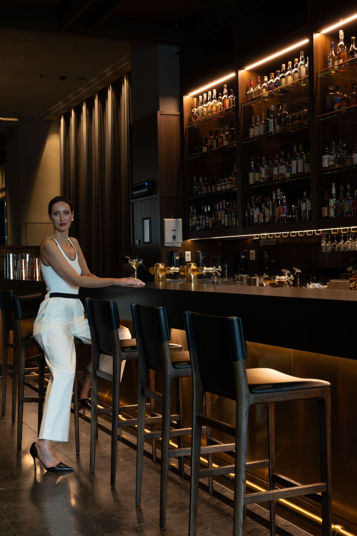Elegant woman in white outfit leaning on bar counter at upscale lounge with backlit liquor shelves.