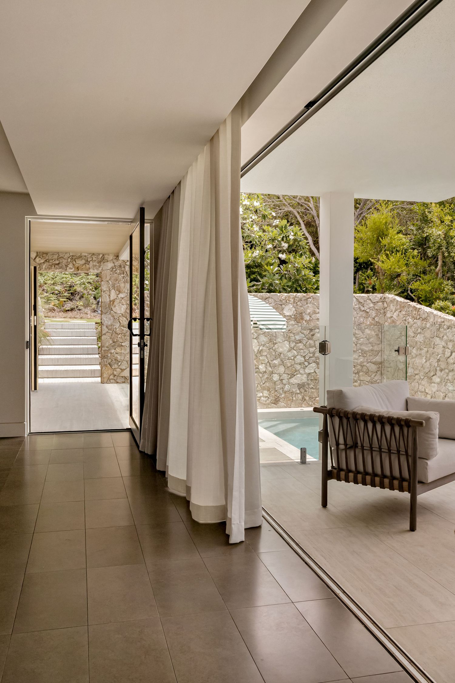 Modern villa interior with sheer white curtains, tiled floors, and open sliding doors leading to a private pool and stone wall garden.