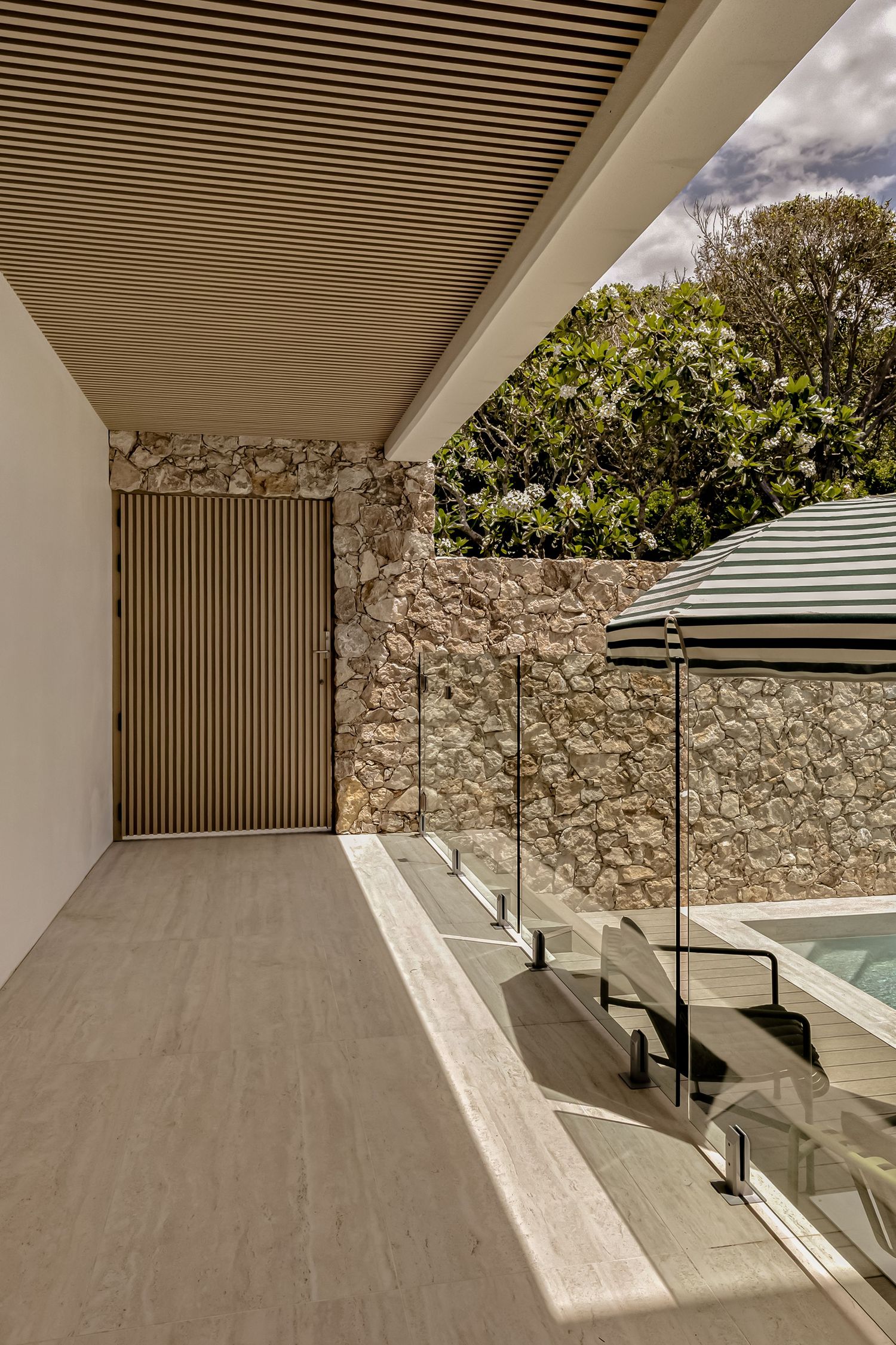 Modern terrace with glass balustrade, stone wall, timber ceiling, and striped umbrella overlooking lush greenery.