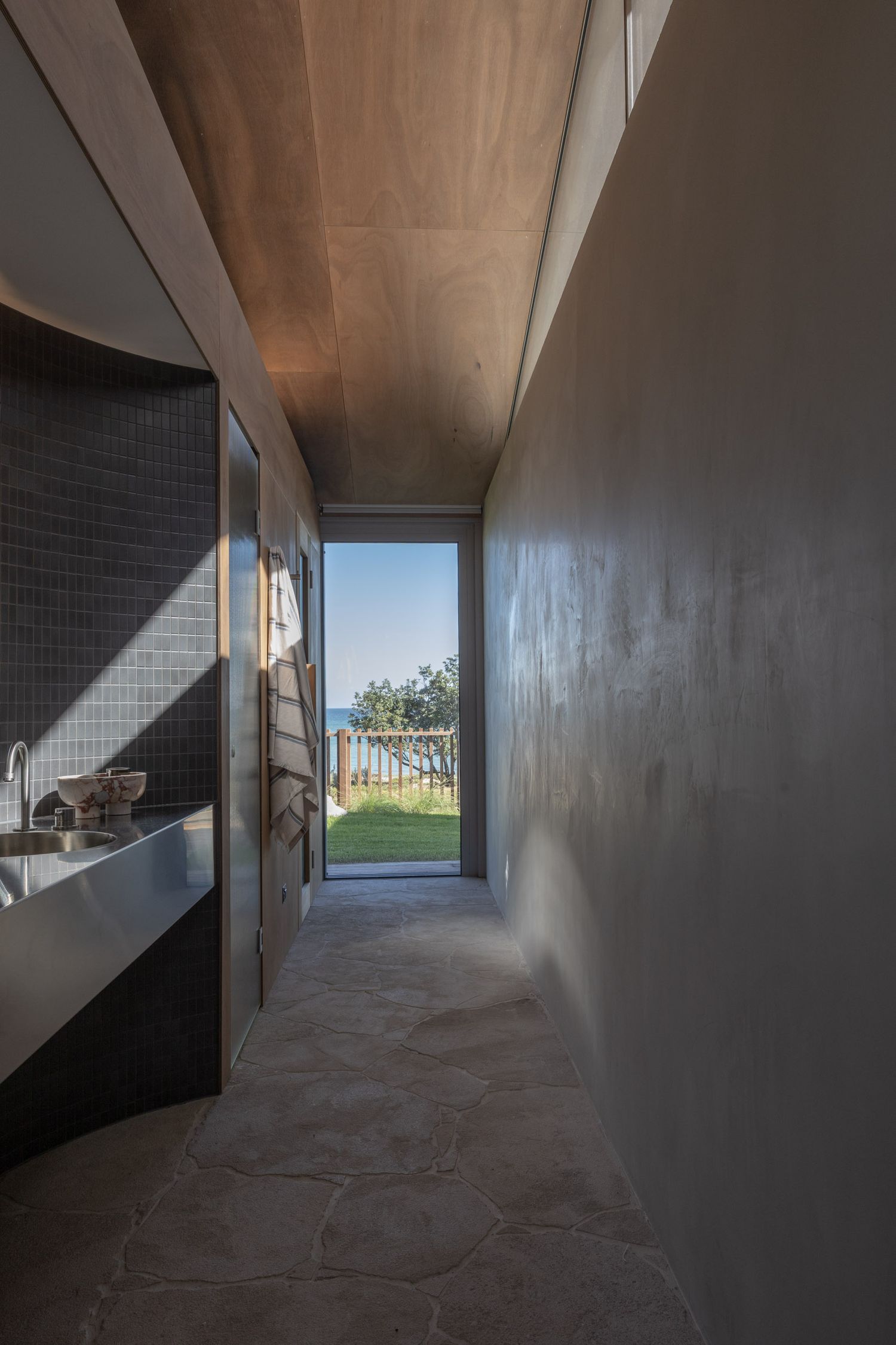 Modern hallway with concrete floors leads to bright window view of ocean and grass through contemporary architectural design.