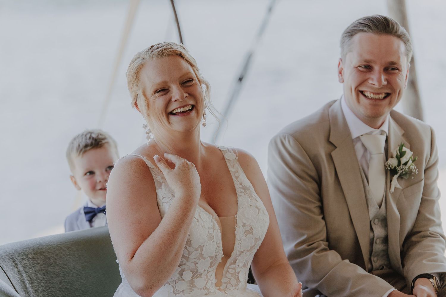 Smiling bride in lace gown and groom in tan suit laugh during wedding ceremony with young boy in bow tie behind them.