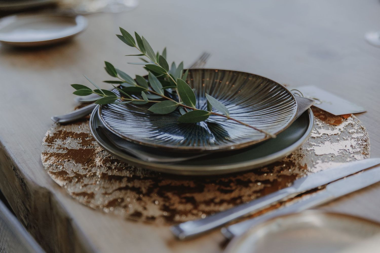 Rustic wedding table setting with dark ceramic plates, olive branch, and gold sequin placemat on a wooden table.