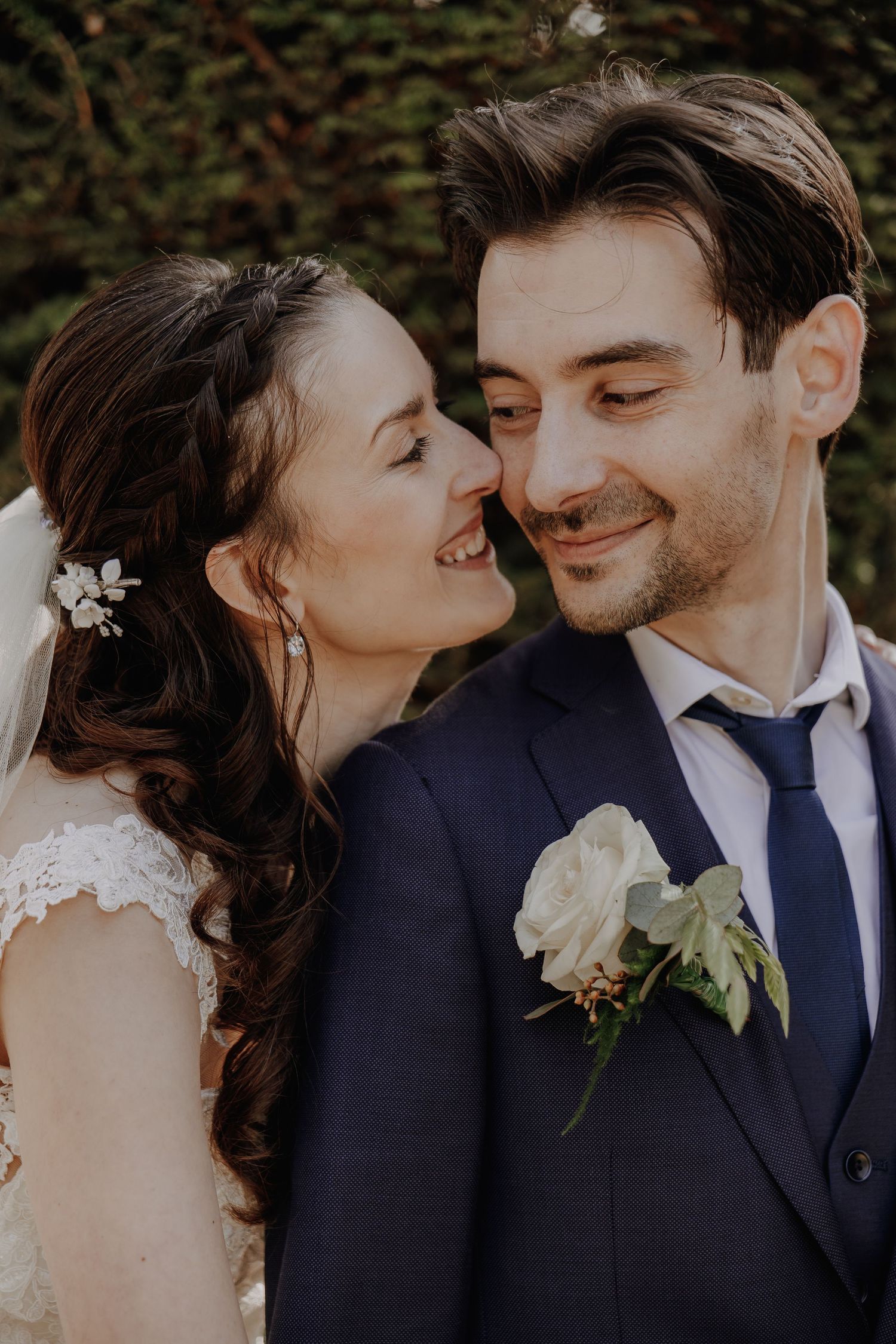 A smiling bride and groom share a tender moment, with the groom wearing a navy suit and white boutonniere.