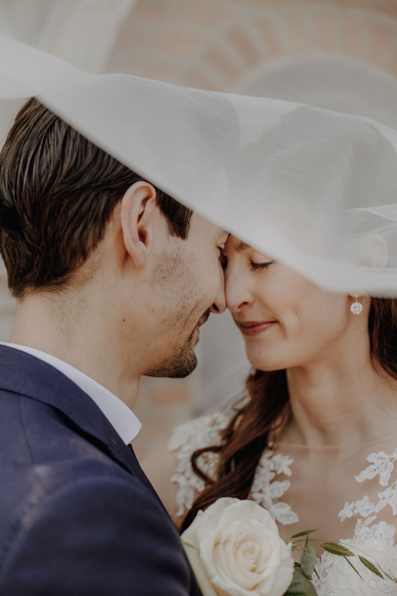Romantic wedding couple touching foreheads under brides flowing veil, bride holding white rose bouquet.