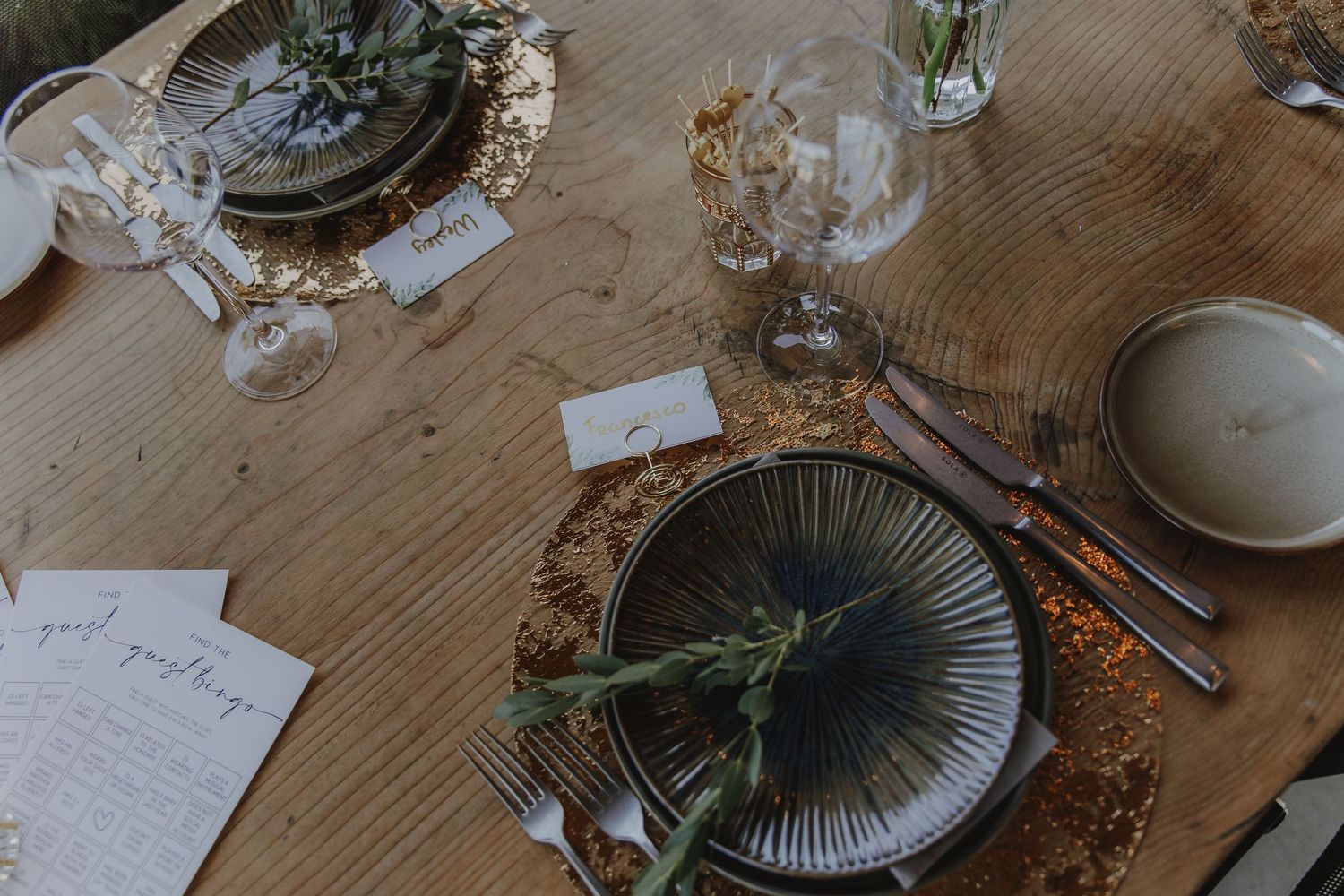 Elegant rustic wedding table setting with dark ceramic plates, greenery, and place cards on wood.