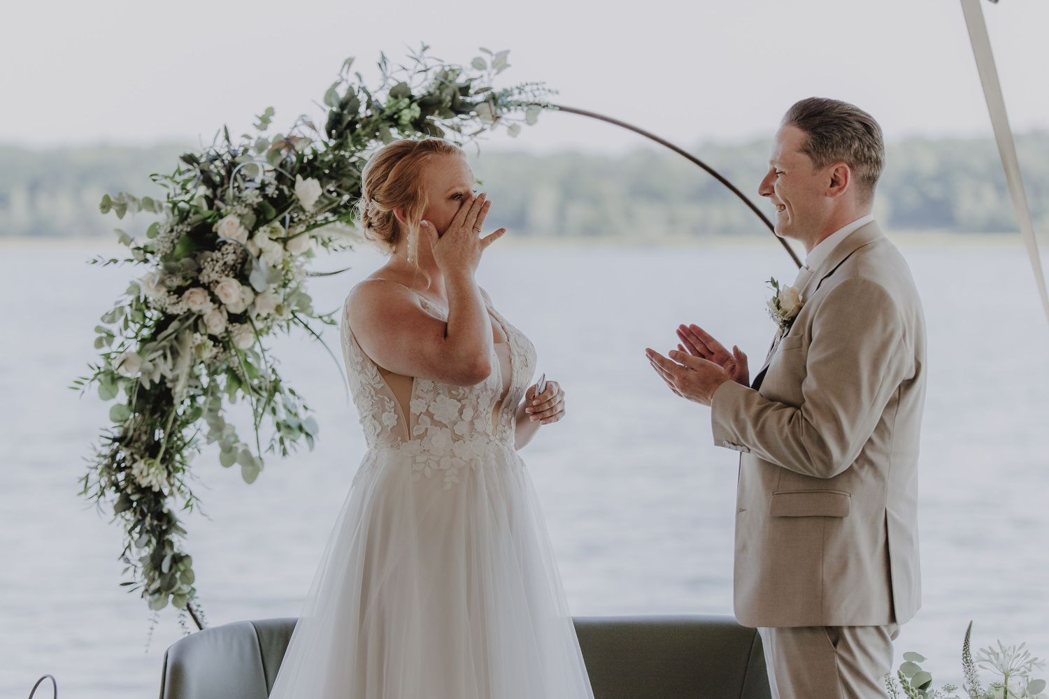 Emotional bride wiping tears at outdoor waterfront wedding ceremony with floral arch, as groom in tan suit applauds.