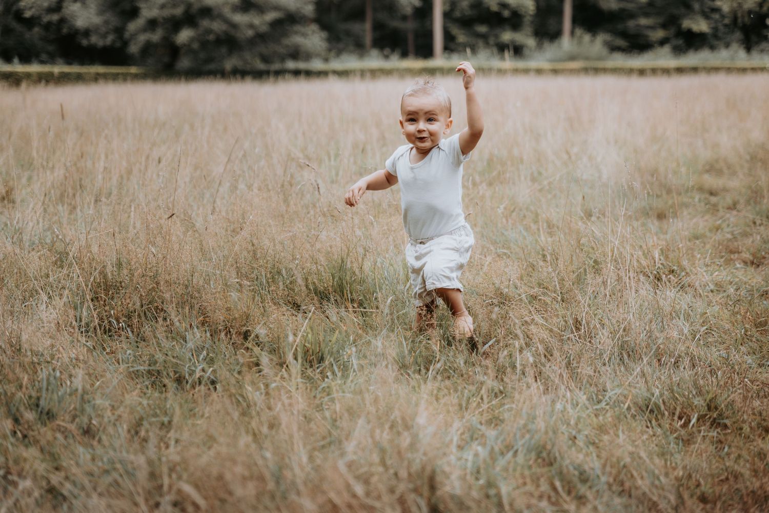 Happy toddler in white outfit runs through a golden grass field outdoors.