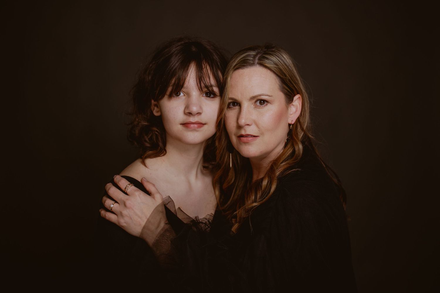 A mother and daughter pose together in a dark studio portrait, both wearing black, sharing a warm embrace.