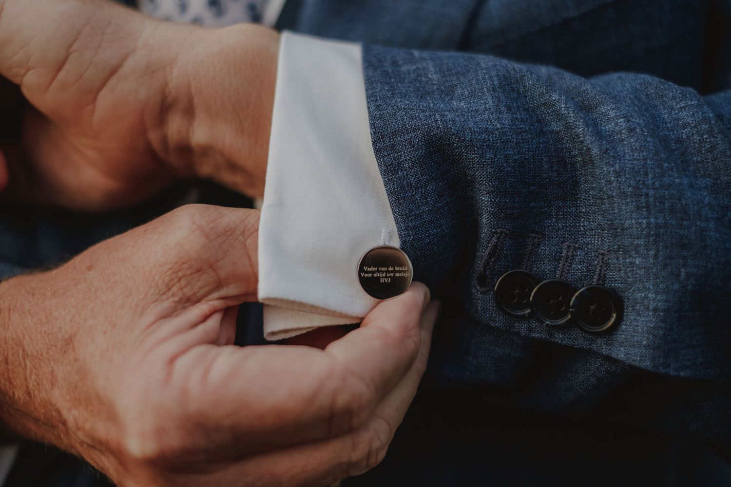Close-up of a man fastening a round engraved cufflink on a white dress shirt with a blue tweed suit jacket.
