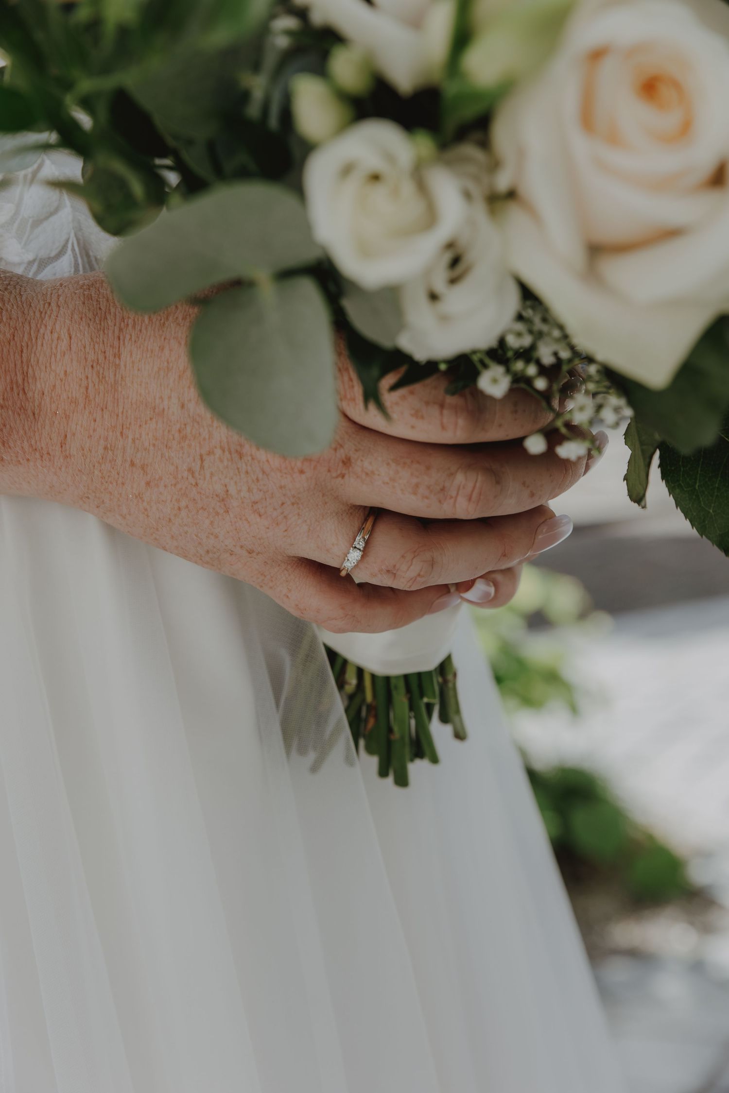 Bride holding a white rose and greenery bouquet with a diamond engagement ring on her finger.