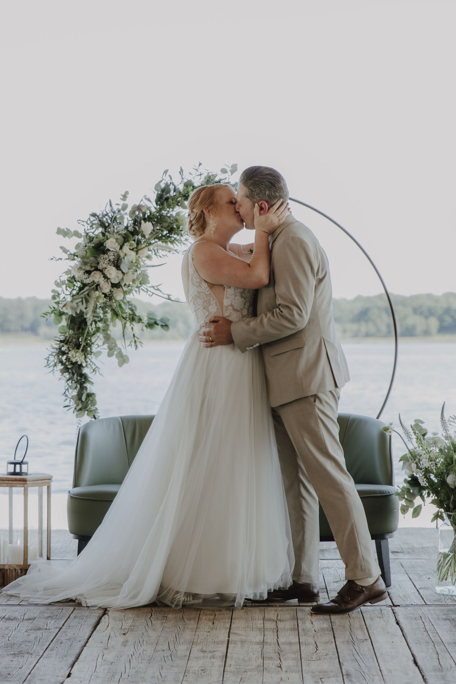 Bride and groom share first kiss at waterfront wedding ceremony beneath circular floral arch with white roses and greenery.