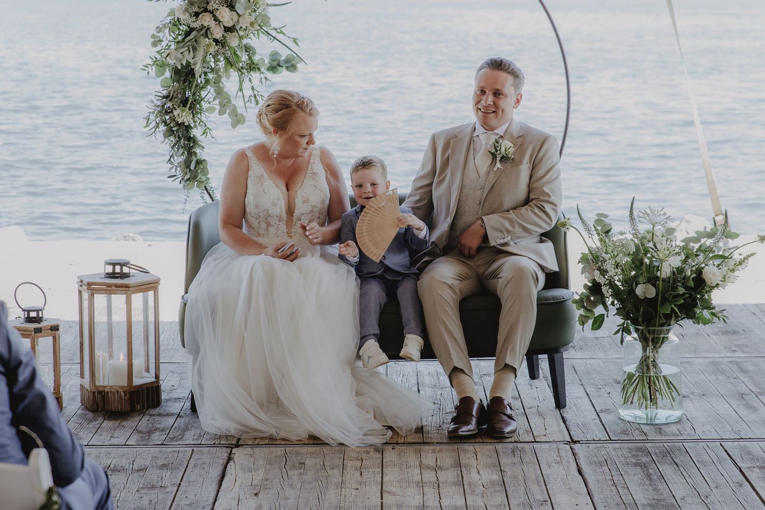 Bride and groom sit with young child during outdoor waterfront wedding ceremony on wooden dock.