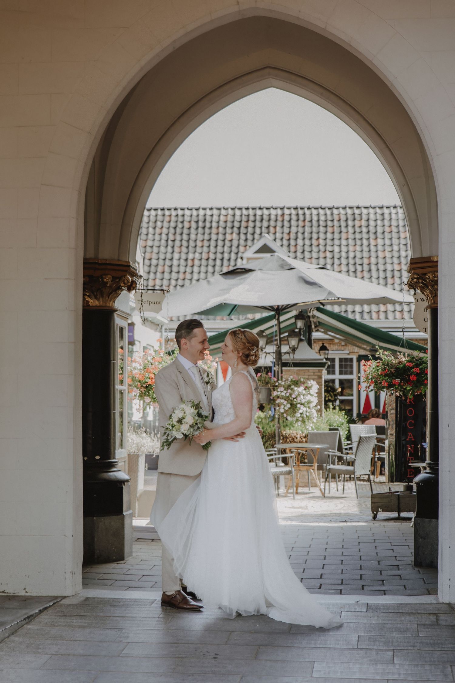 Bride and groom embrace under gothic stone arch at outdoor wedding venue with garden patio in background.