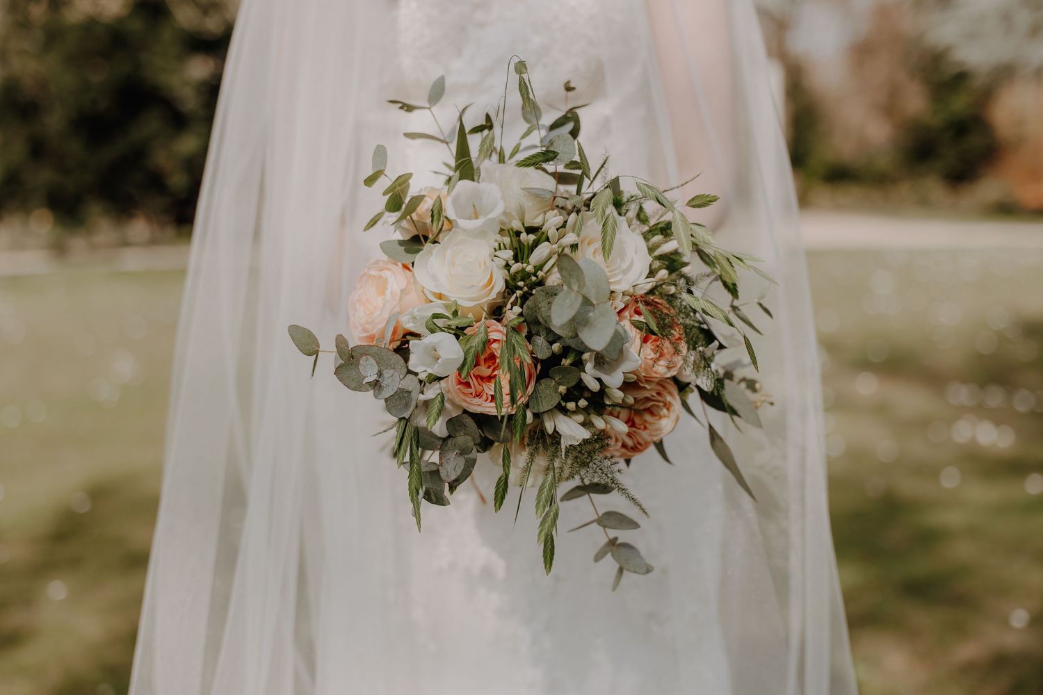 Bride holding a lush wedding bouquet with white and peach roses, eucalyptus greenery, wearing a white gown and veil.