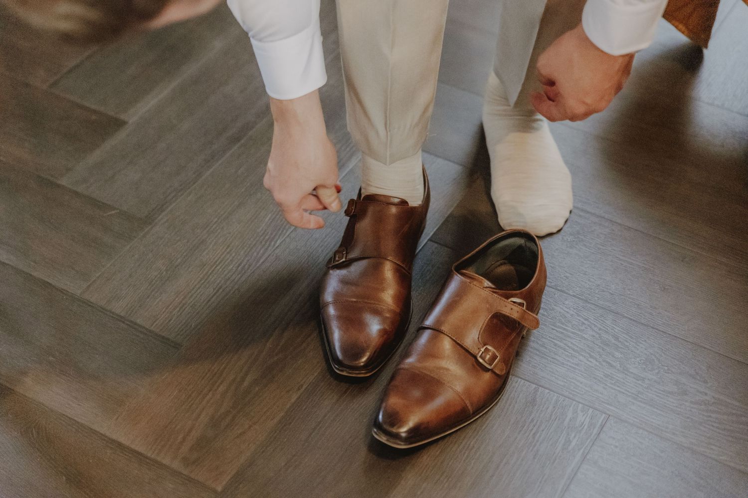 Man in white pants putting on brown leather monk strap dress shoes on herringbone floor.