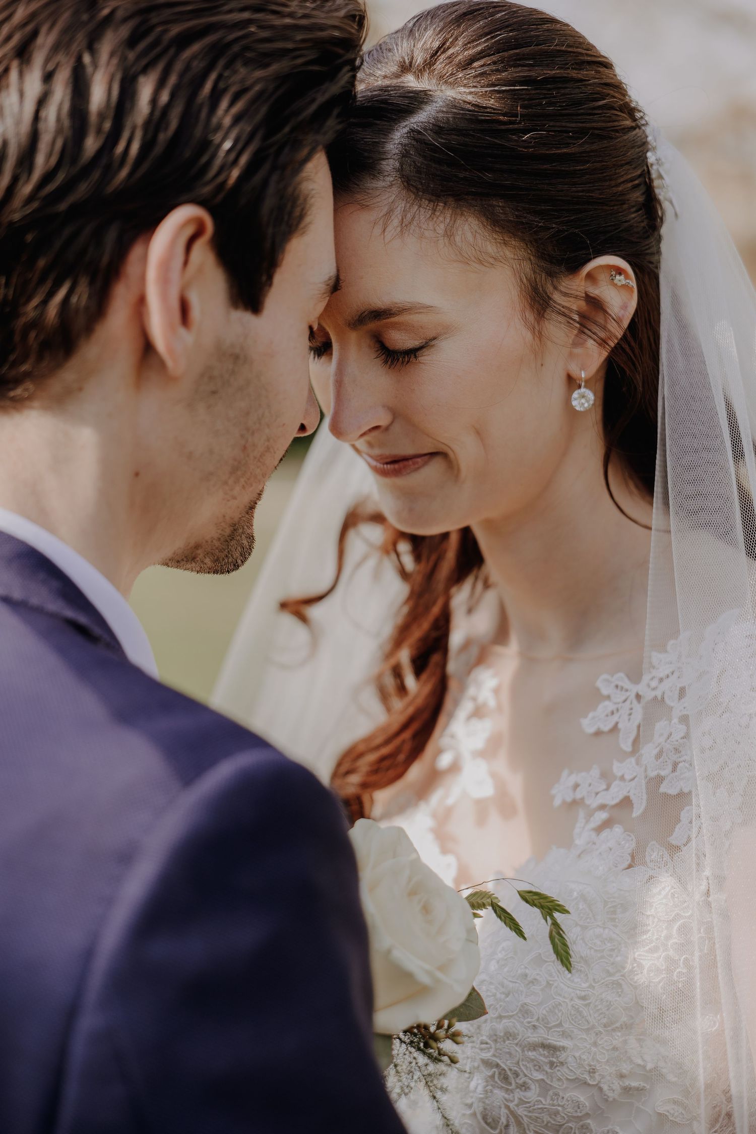 Bride and groom touching foreheads tenderly, bride wearing lace dress and veil with pearl earrings.