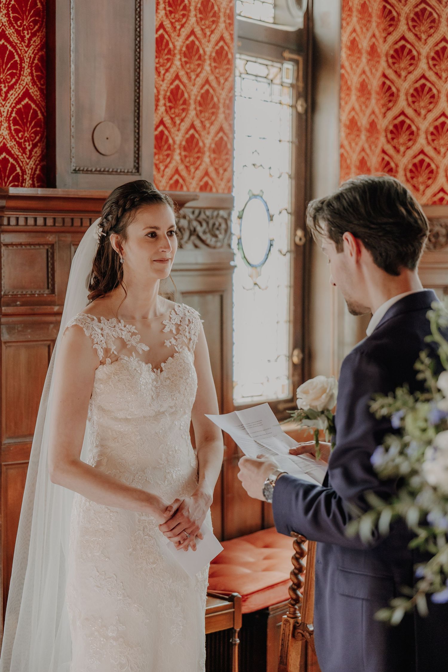Bride in lace wedding dress smiling at groom reading vows in ornate red room.