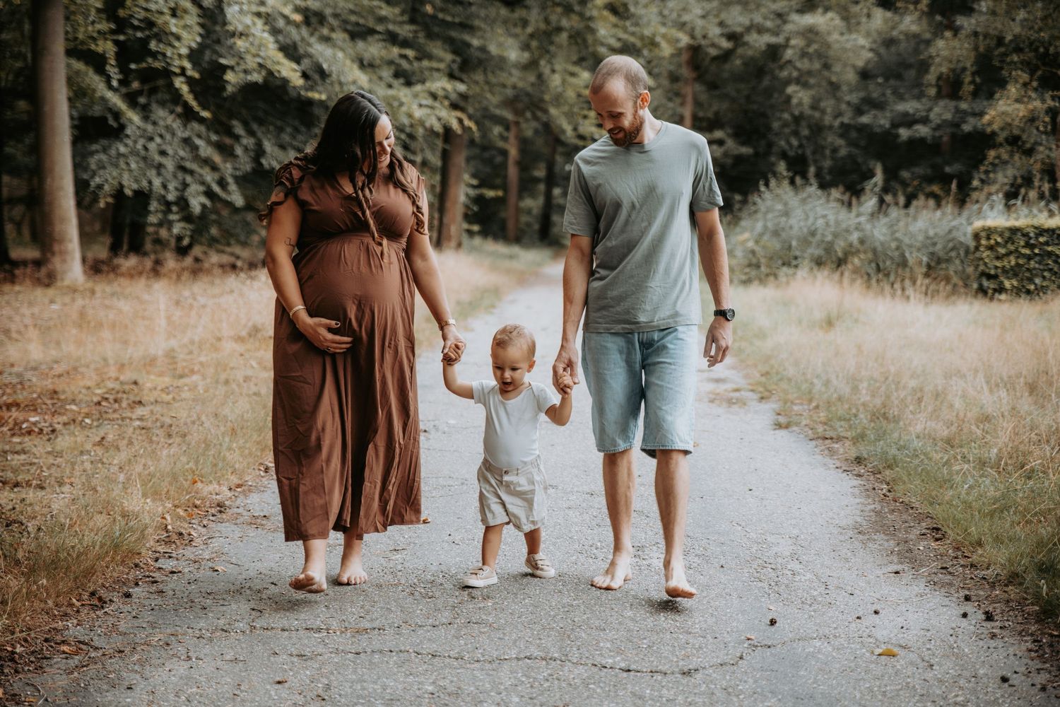 Pregnant woman in brown dress walks with partner and toddler on a sunny park path.