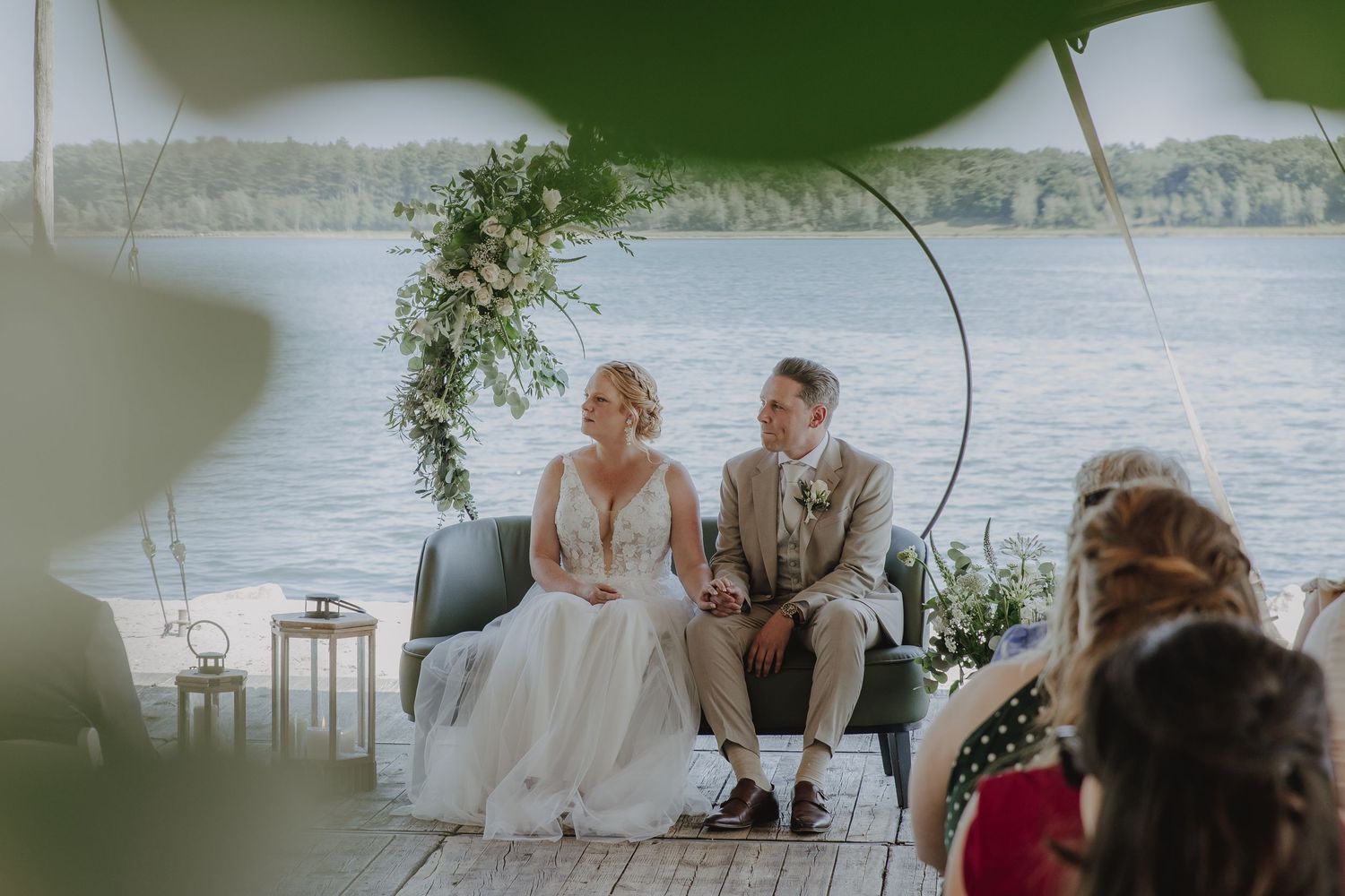 Bride and groom sit together on a sofa during outdoor waterfront wedding ceremony with floral arch backdrop.