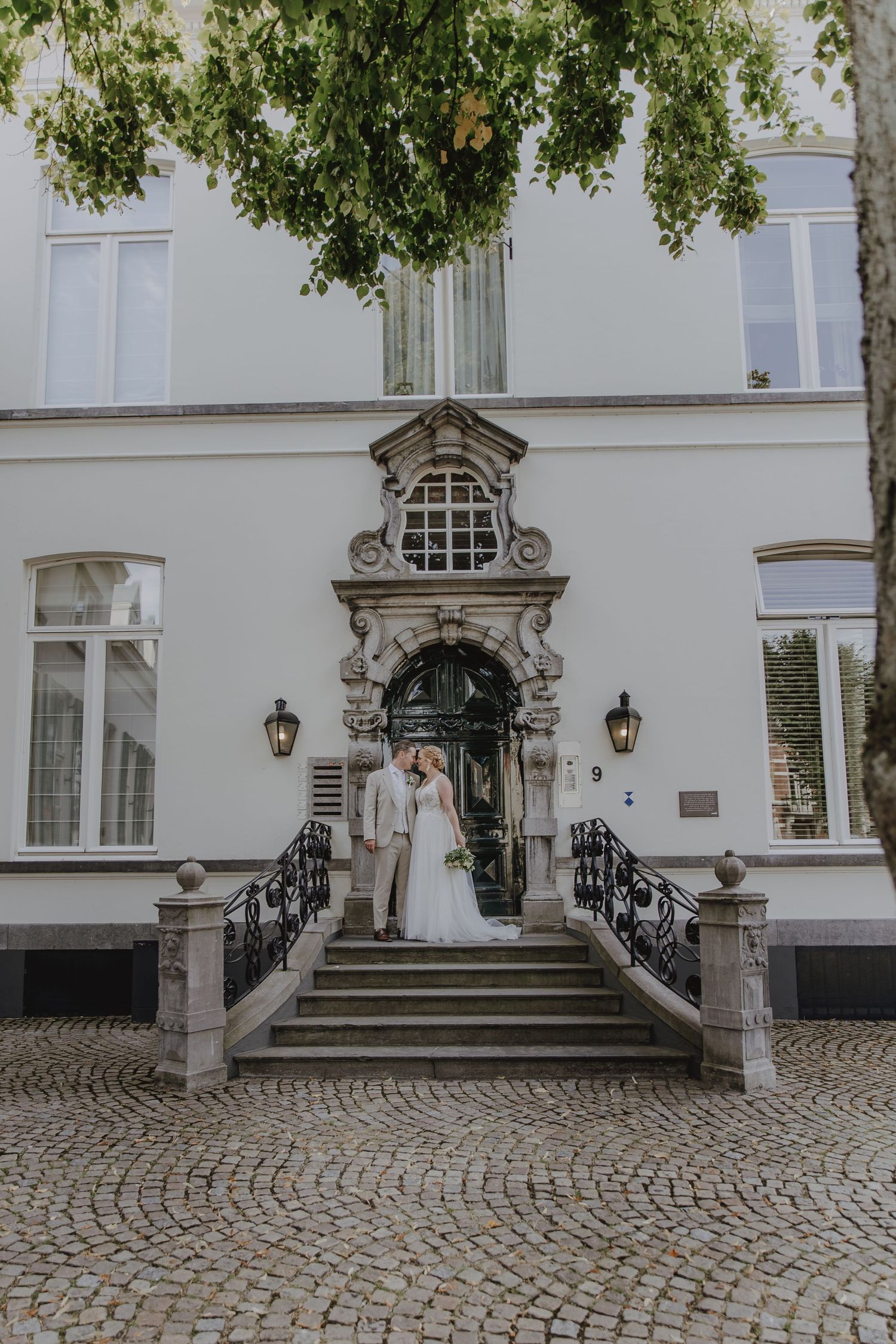 Wedding couple poses on ornate stone staircase entrance of elegant white European manor building.