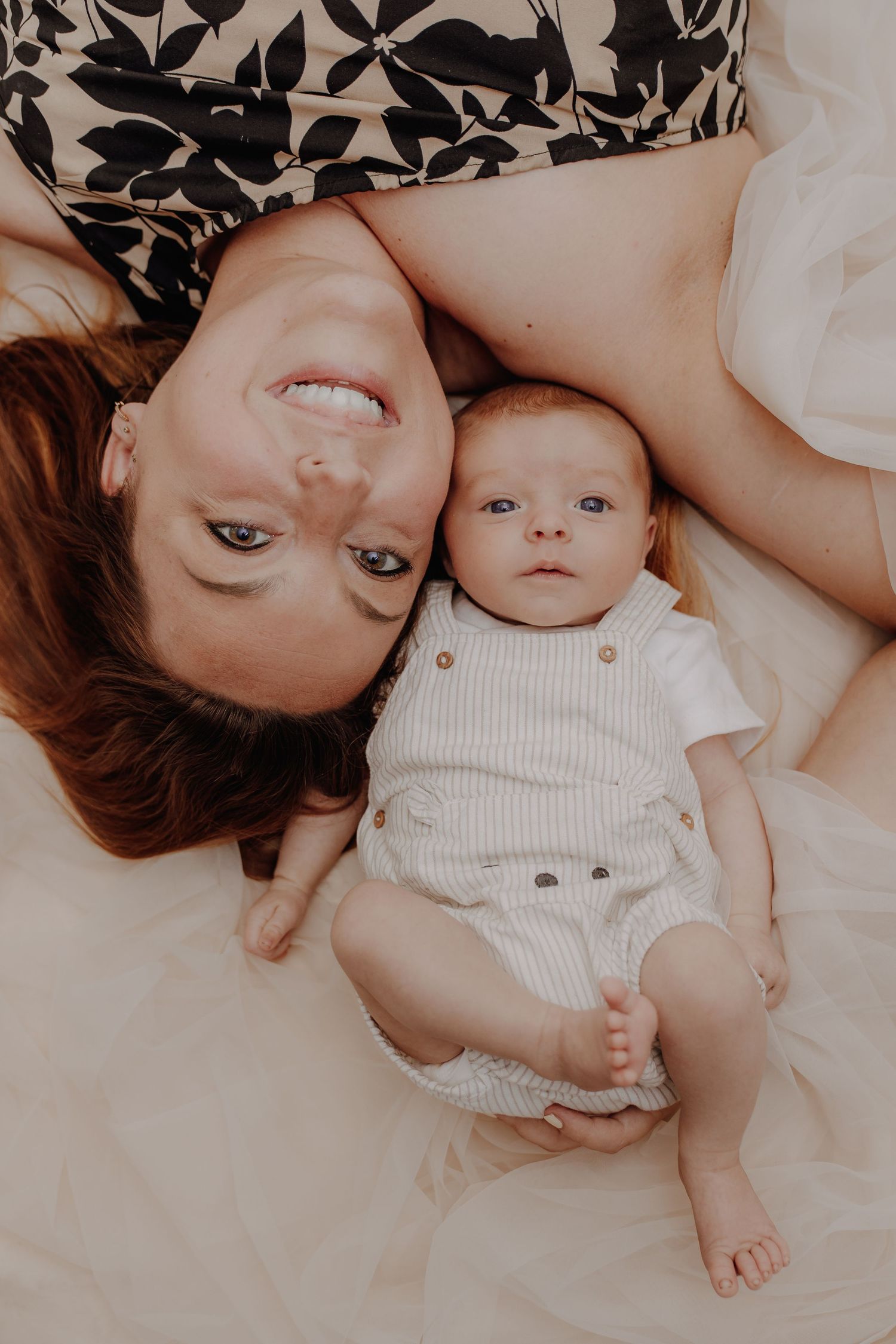 A smiling mother with red hair lies upside down beside her newborn baby in a white outfit on a cream bed.