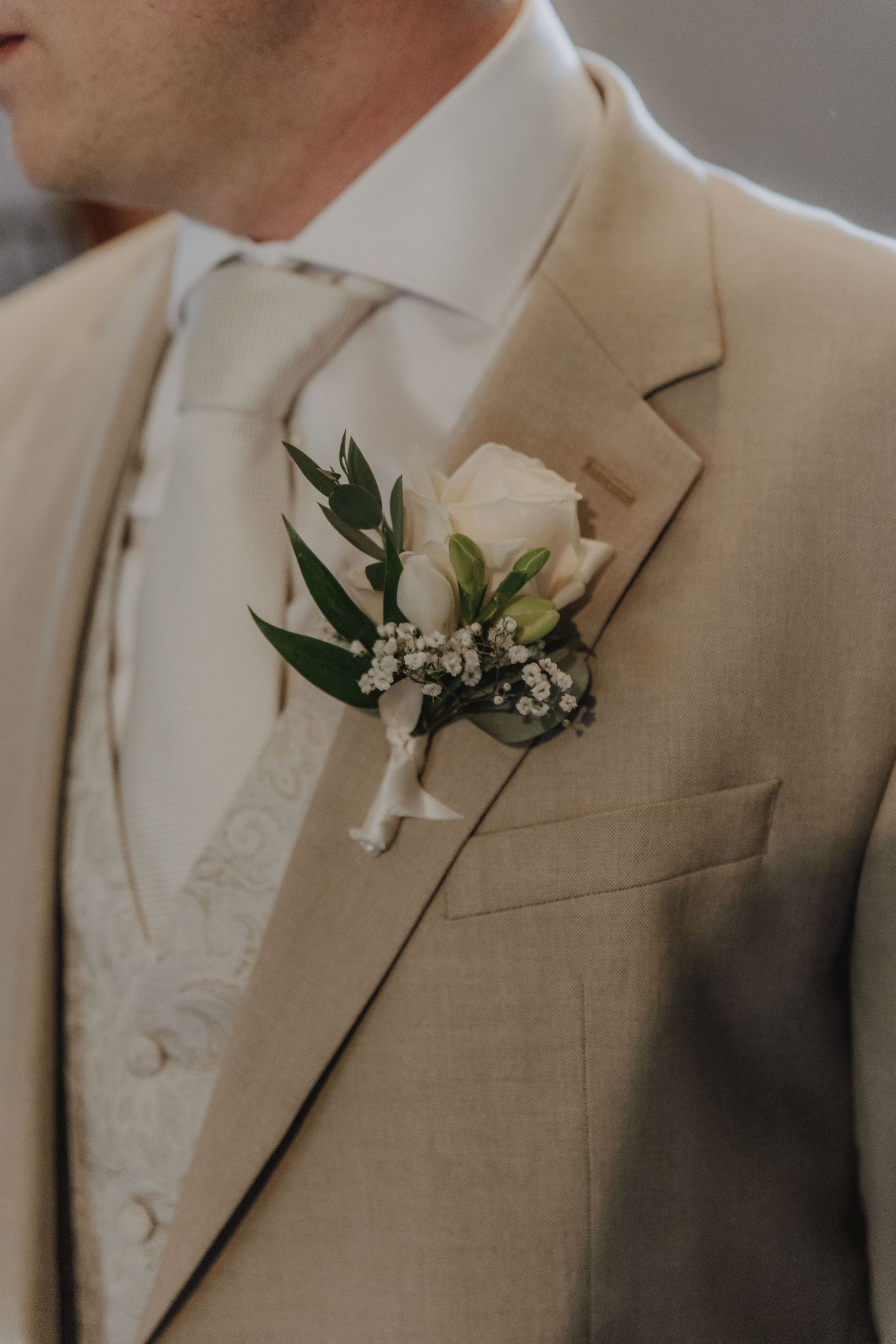Groom wearing beige suit with white rose boutonniere and cream tie at wedding.