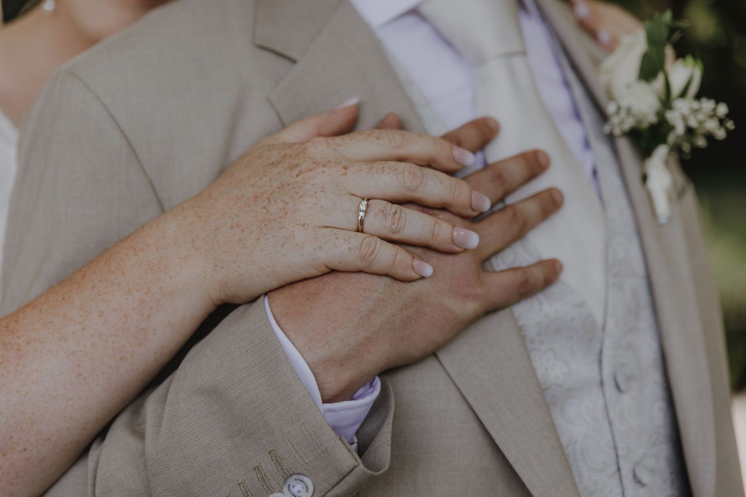 Bride and groom showing wedding rings with hands on grooms beige suit chest.