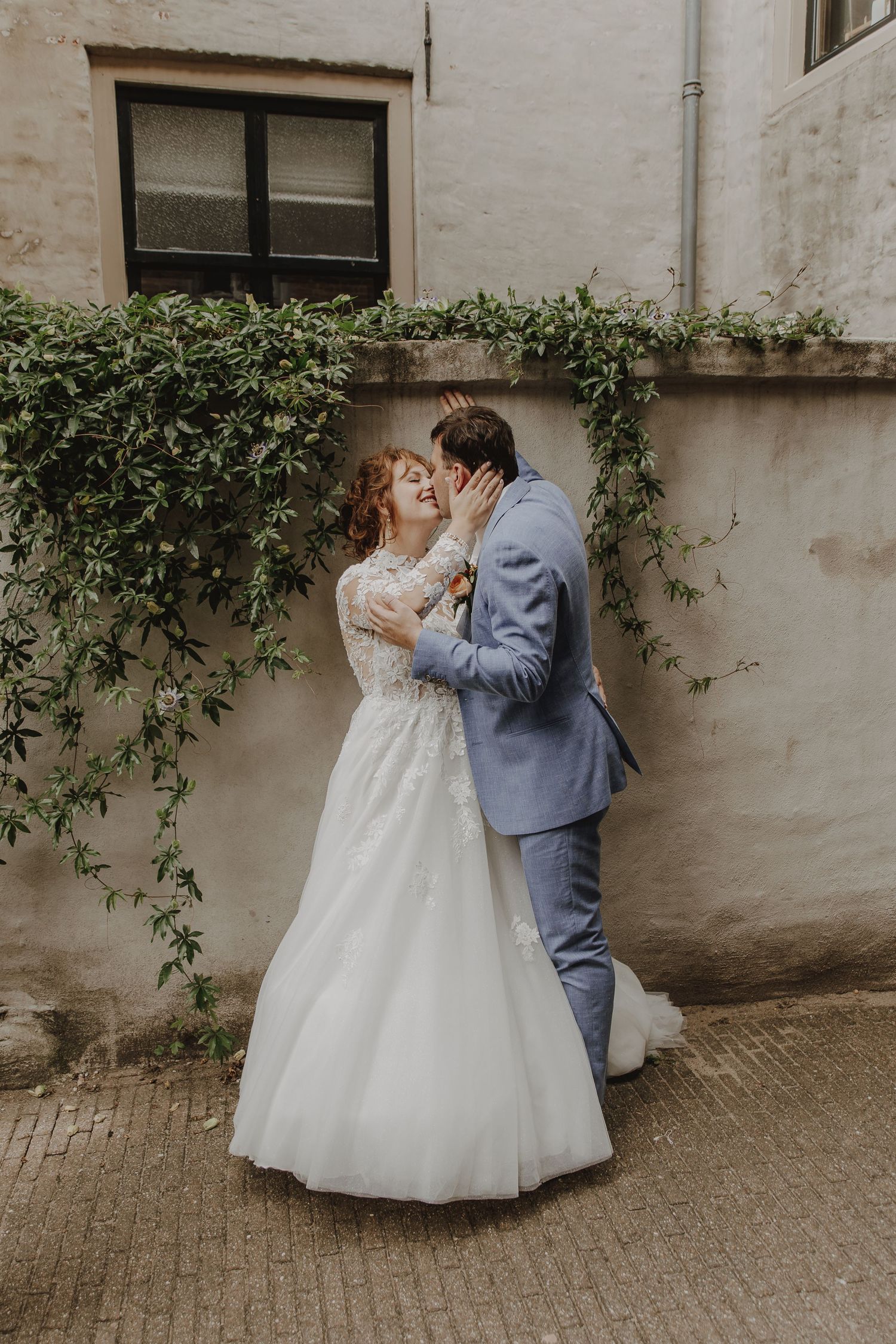 A bride and groom share a romantic kiss outdoors against a stone wall with lush ivy, bride in white gown, groom in blue suit.