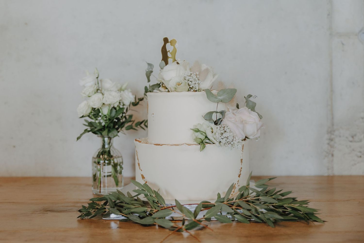 Two-tier white wedding cake with gold trim, floral topper, white peonies, and eucalyptus greenery on wooden table.